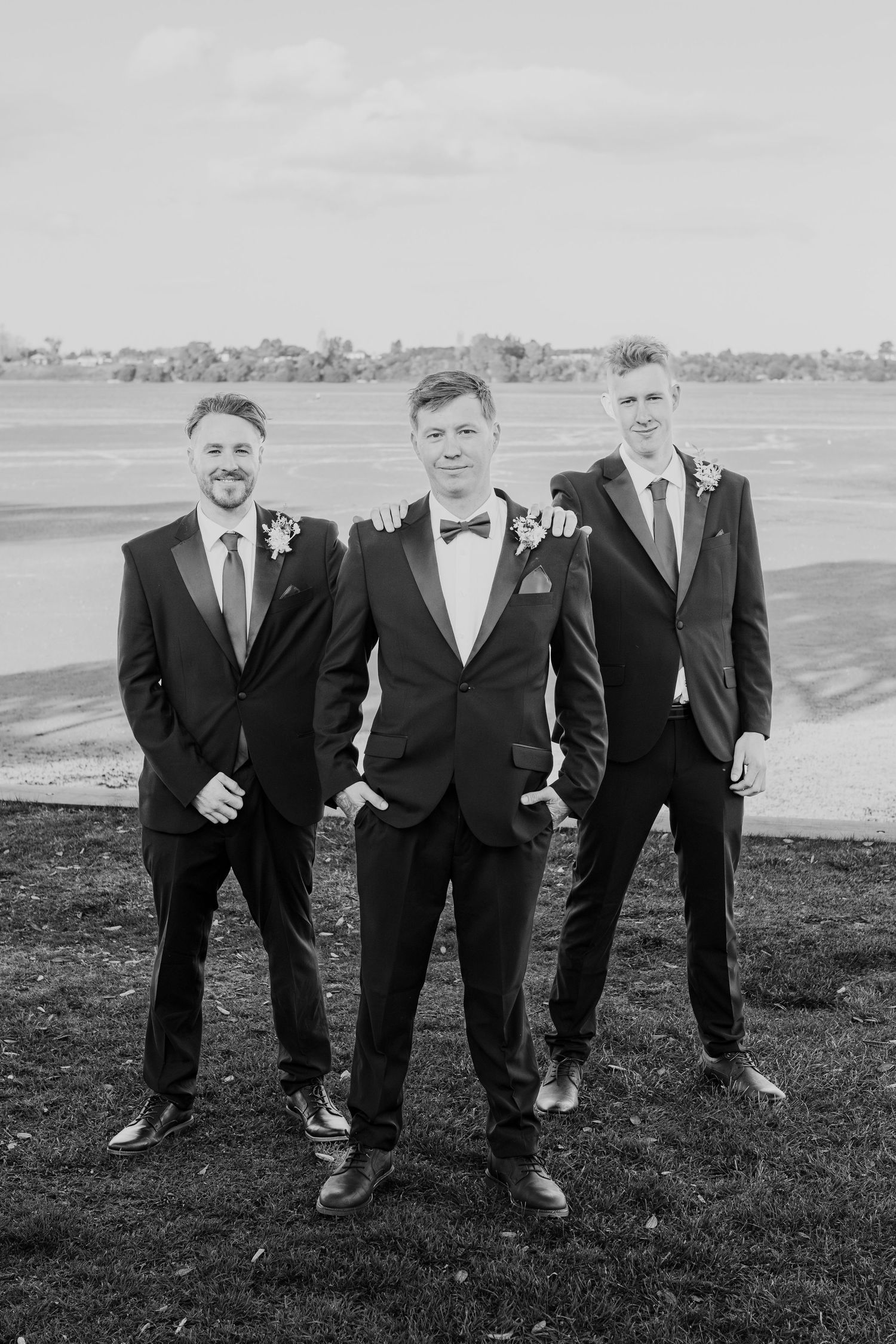Three men in formal suits and bowties pose for a wedding photo on a grassy hillside.