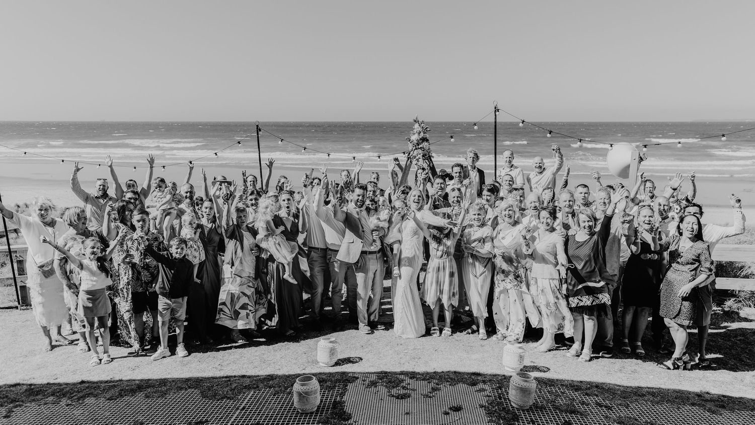 Wedding guests raise their arms in celebration while gathered on a beach for a group photo. Papamoa Surf Lifesaving Club Wedding