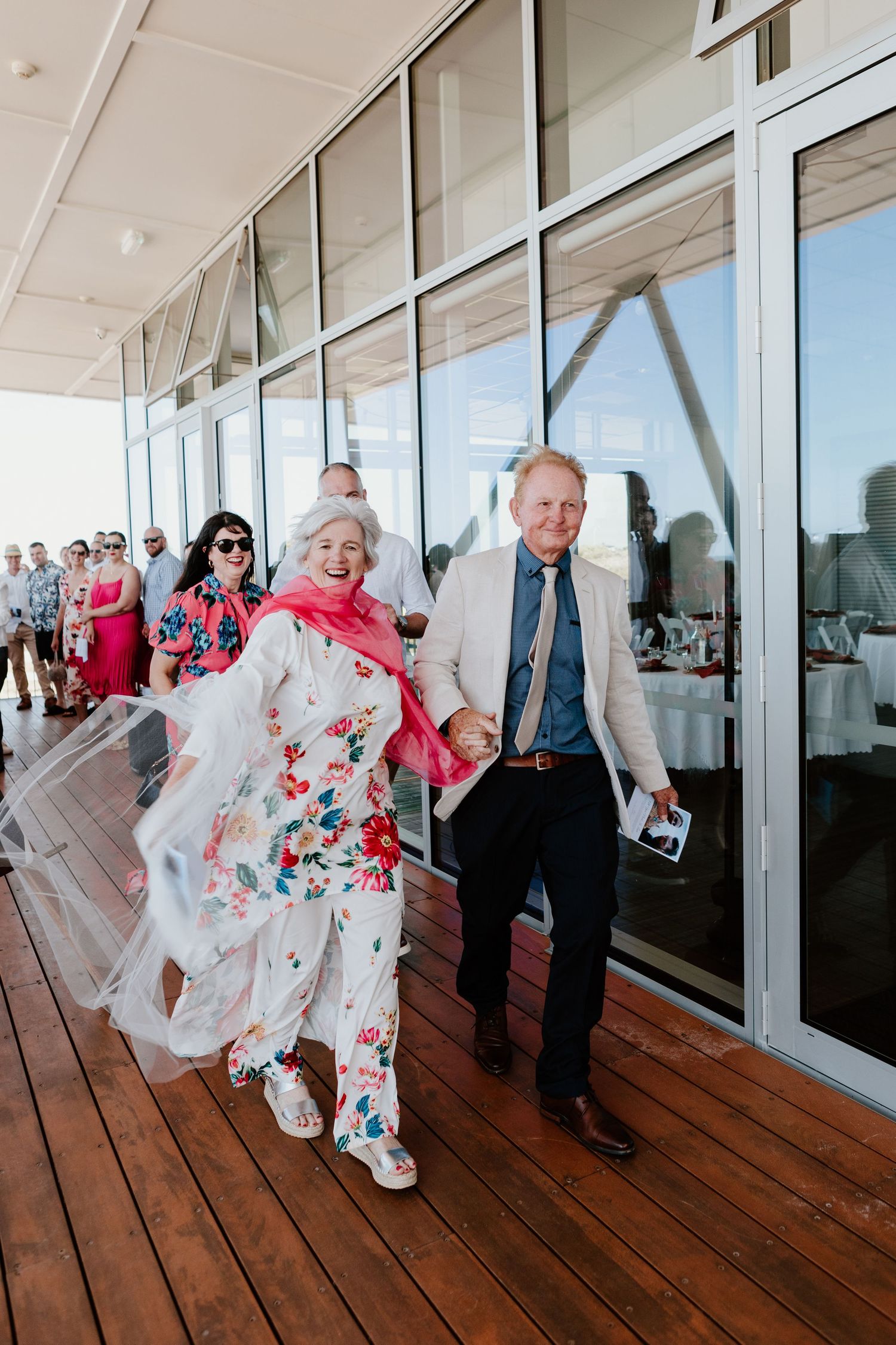 Wedding guests in formal attire dance along a wooden deck with glass windows. Papamoa Surf Lifesaving Club Wedding