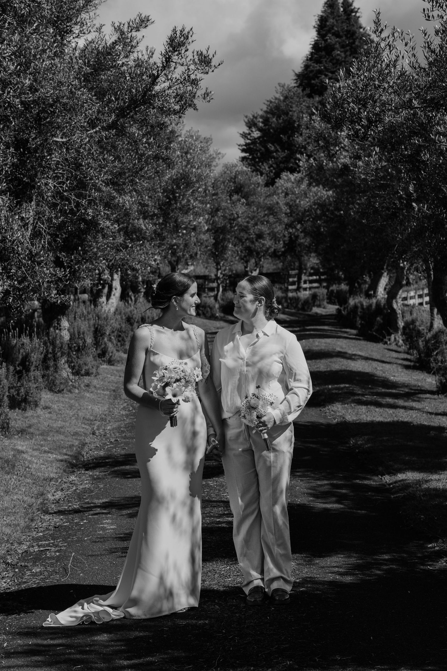 Black and white photo of a bride and groom walking along a tree-lined path, exchanging glances on their wedding day.