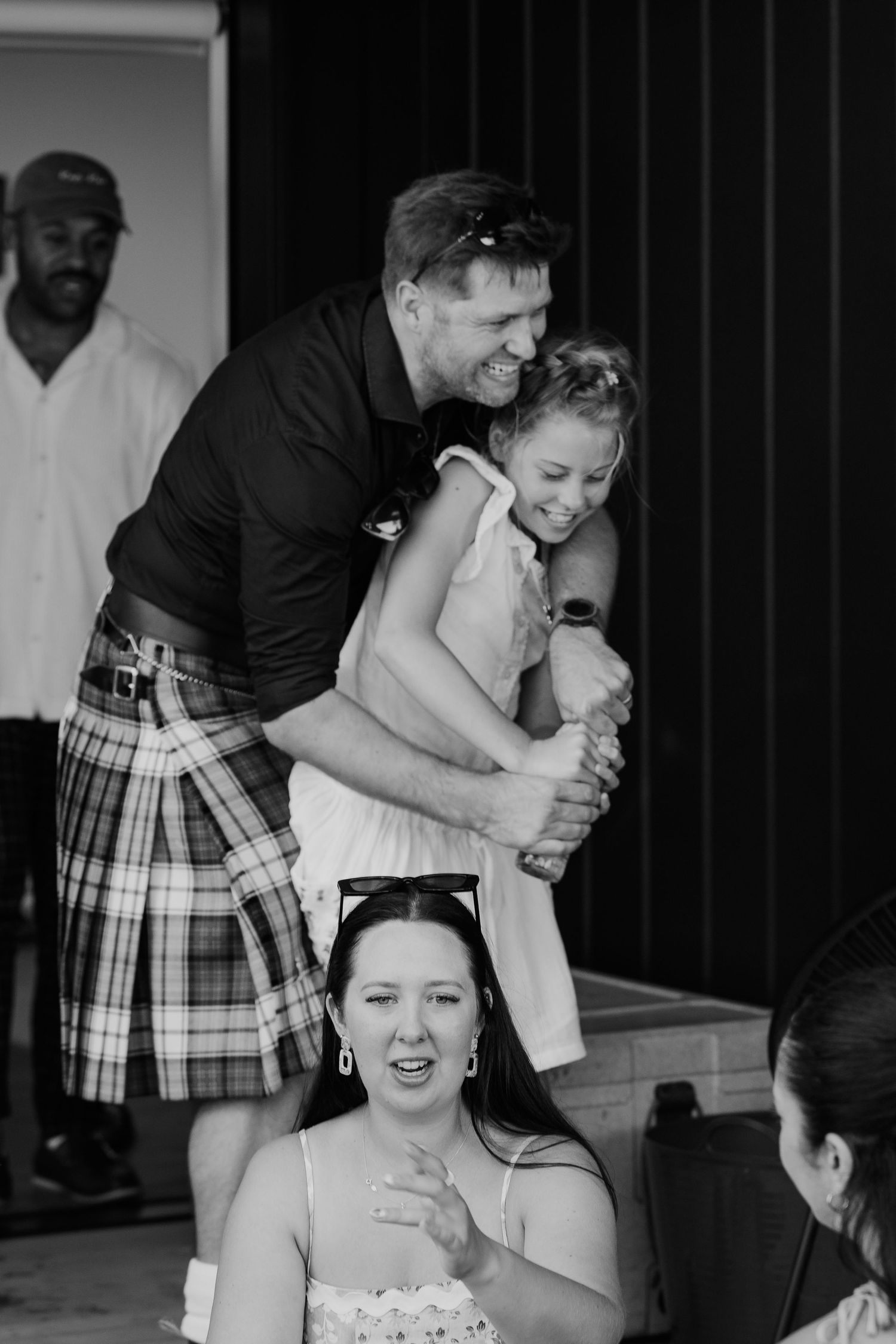 Man in kilt hugging young girl at wedding celebration, woman smiling in foreground, black and white photo.
