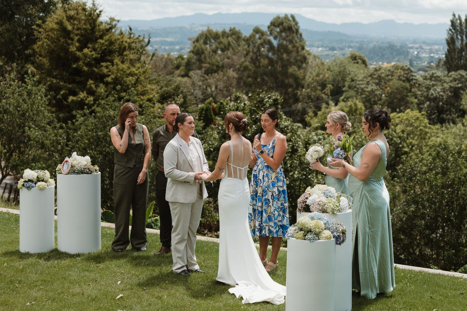 Outdoor wedding ceremony on a hilltop lawn with bride, groom, and guests surrounded by lush greenery and mountain views.