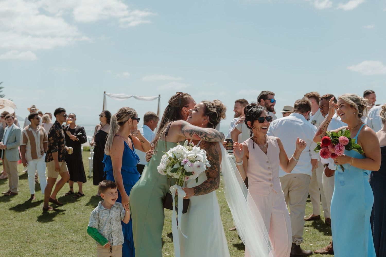 Bride in white gown receives congratulatory hugs from guests at an outdoor wedding ceremony on a sunny day.