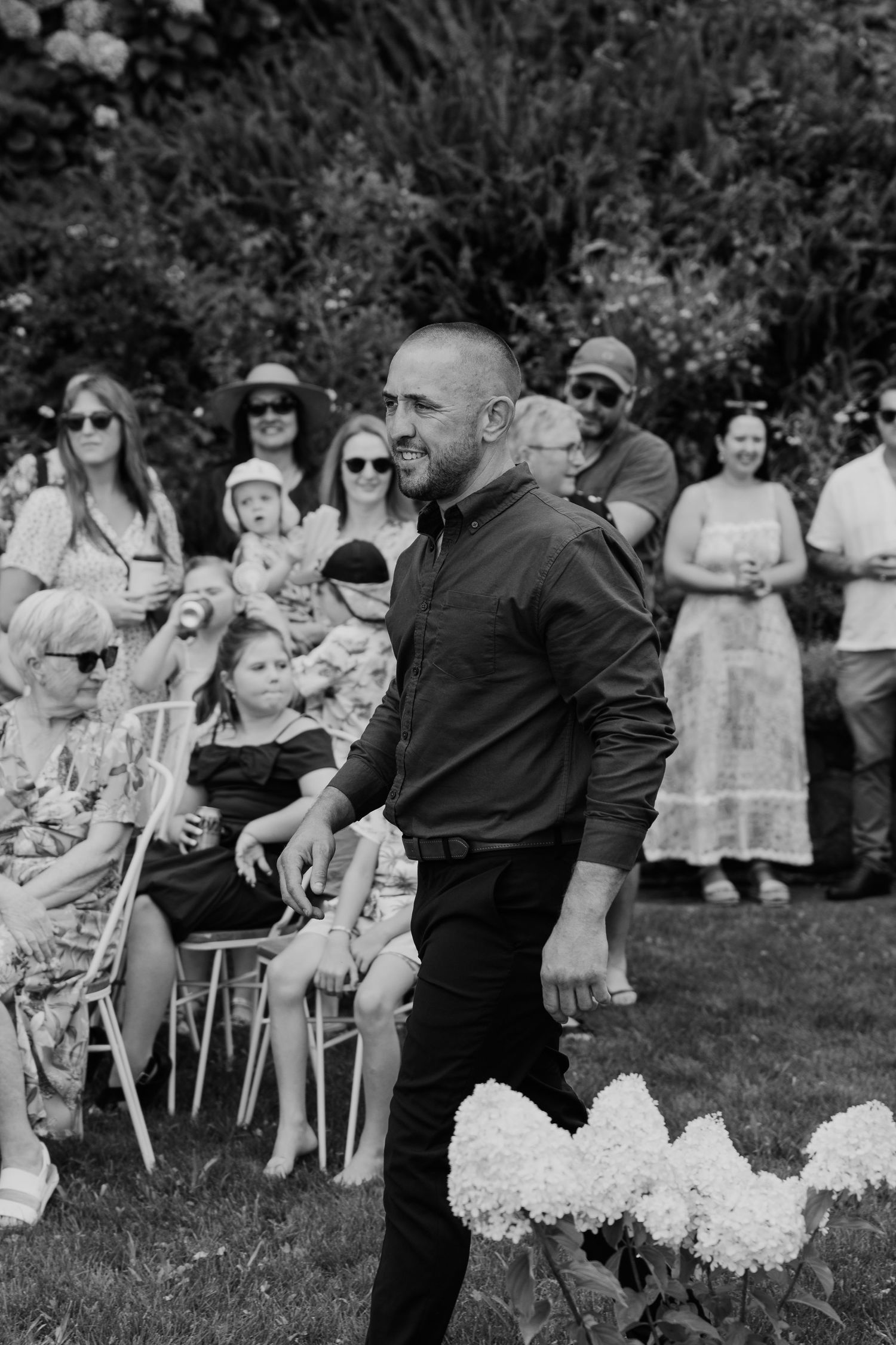 Black and white photo of a bald man in dark clothing walking at an outdoor wedding ceremony with seated guests.