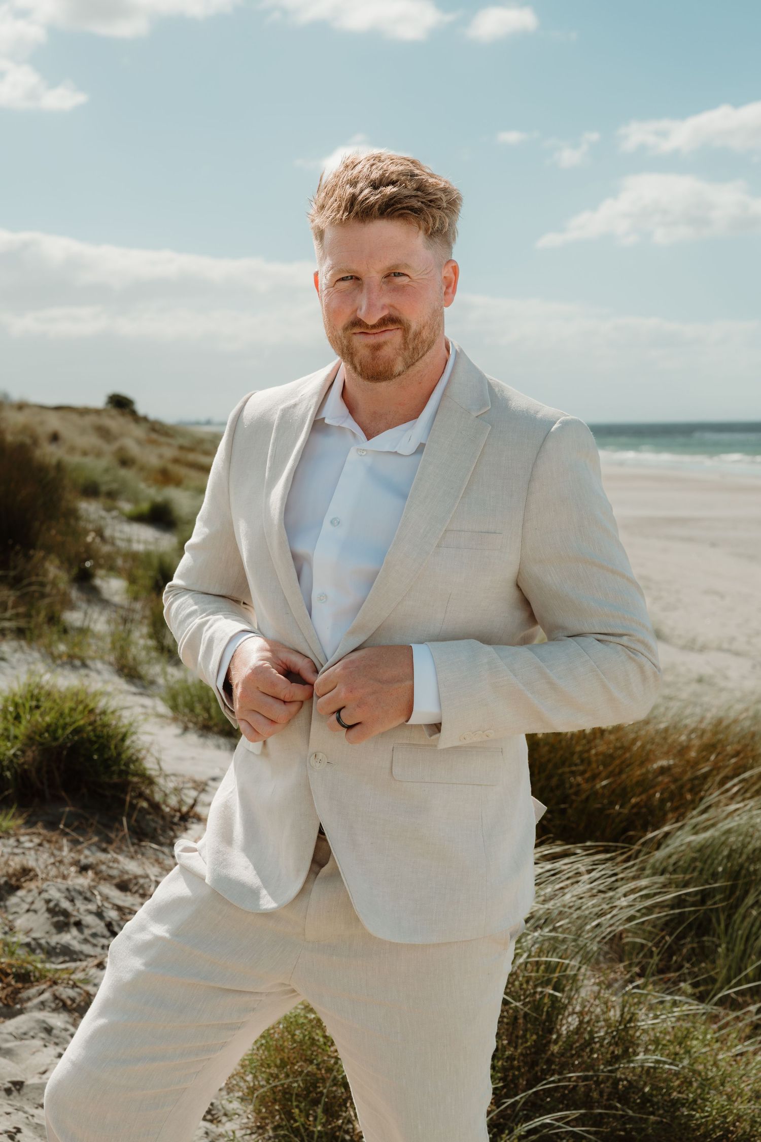 Man in beige linen suit smiling on sandy beach with ocean backdrop on a sunny day.