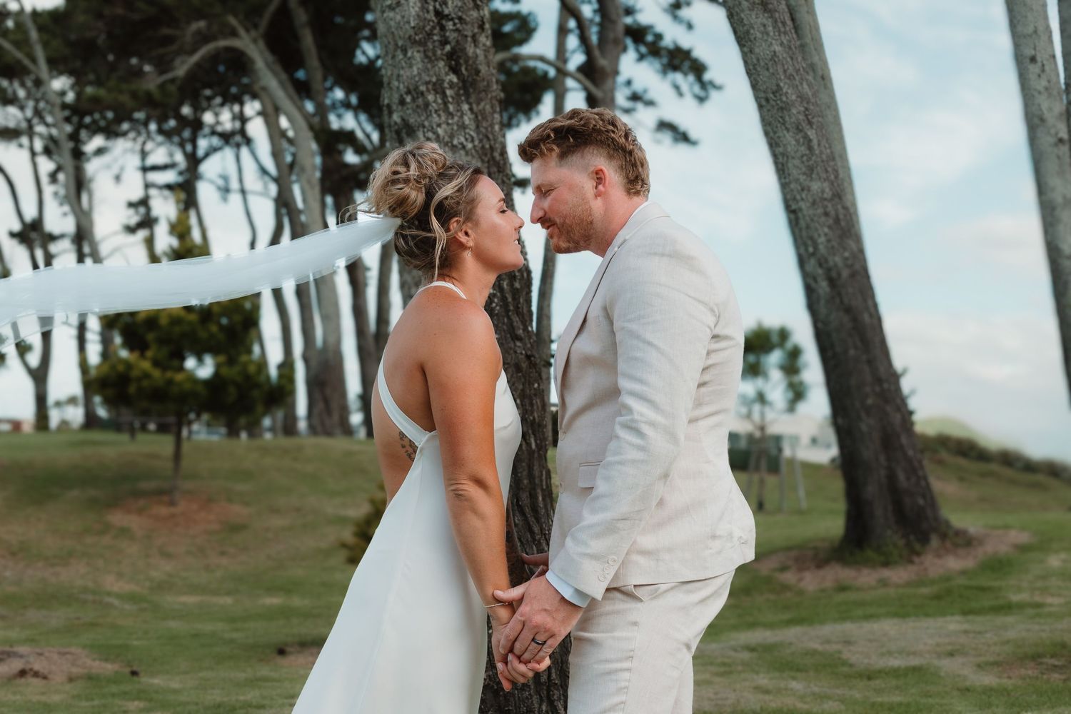Bride and groom holding hands and gazing at each other outdoors near trees on a sunny wedding day.