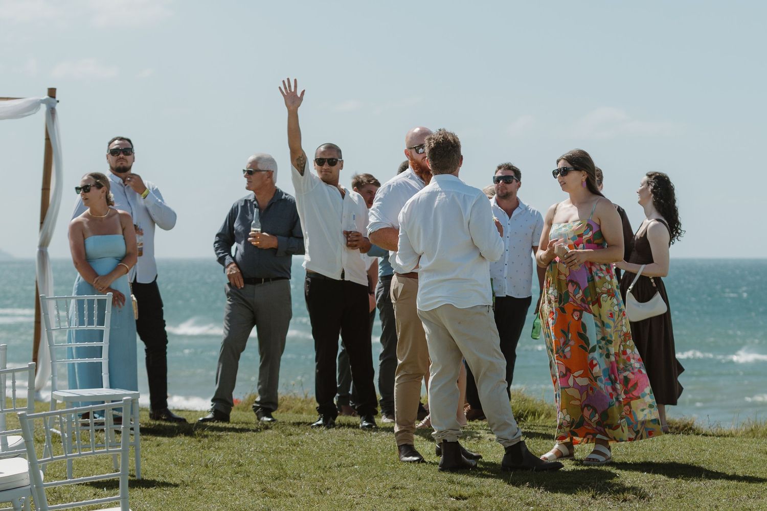 Guests mingle at an outdoor oceanside wedding ceremony on a sunny day, with one person waving their hand.