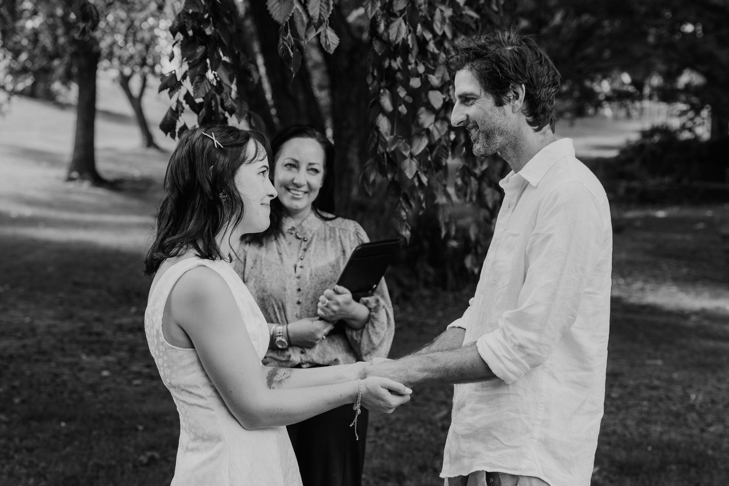 Couple holding hands during outdoor wedding ceremony with officiant in black and white park photo.