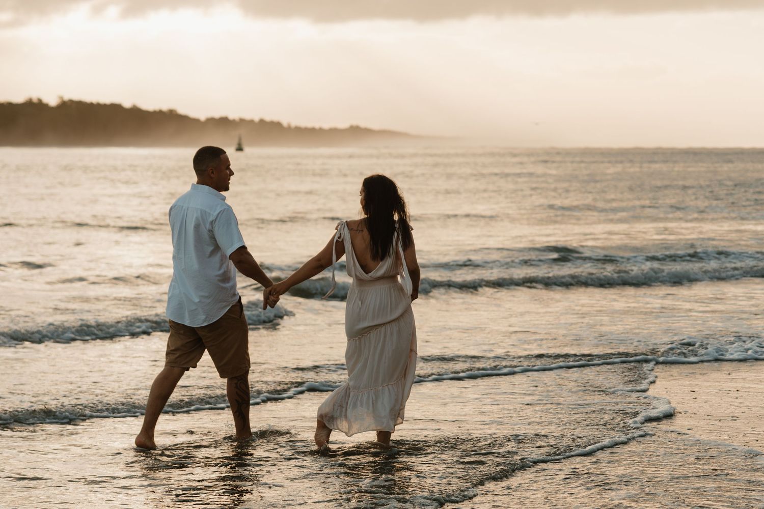 Couple walking together along beach shoreline at sunset holding hands in romantic coastal setting.