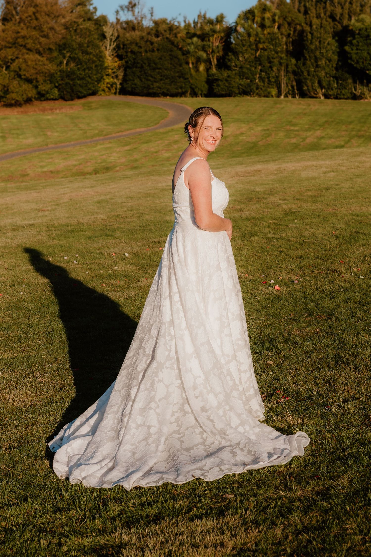 A bride in a flowing white lace wedding gown poses outdoors in sunset light while showing off the dress's elegant train.