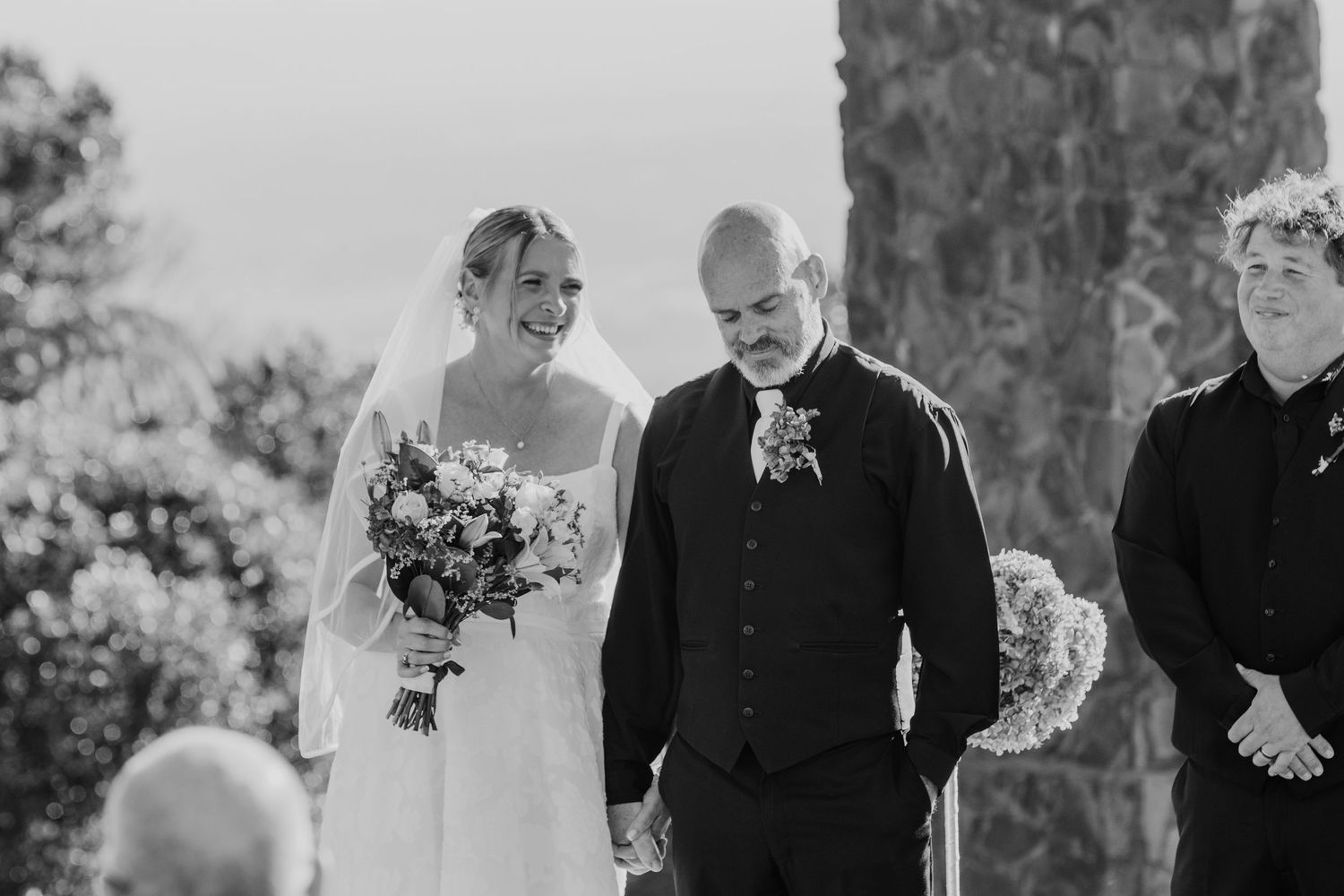 A black and white wedding ceremony photo showing a couple and officiant during an outdoor ceremony at a stone venue.