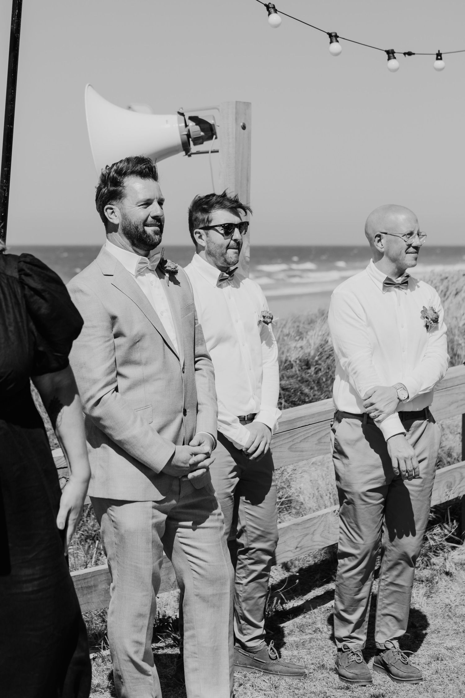Black and white photo of groomsmen wearing light colored suits and white shirts standing together outdoors at a beach wedding.