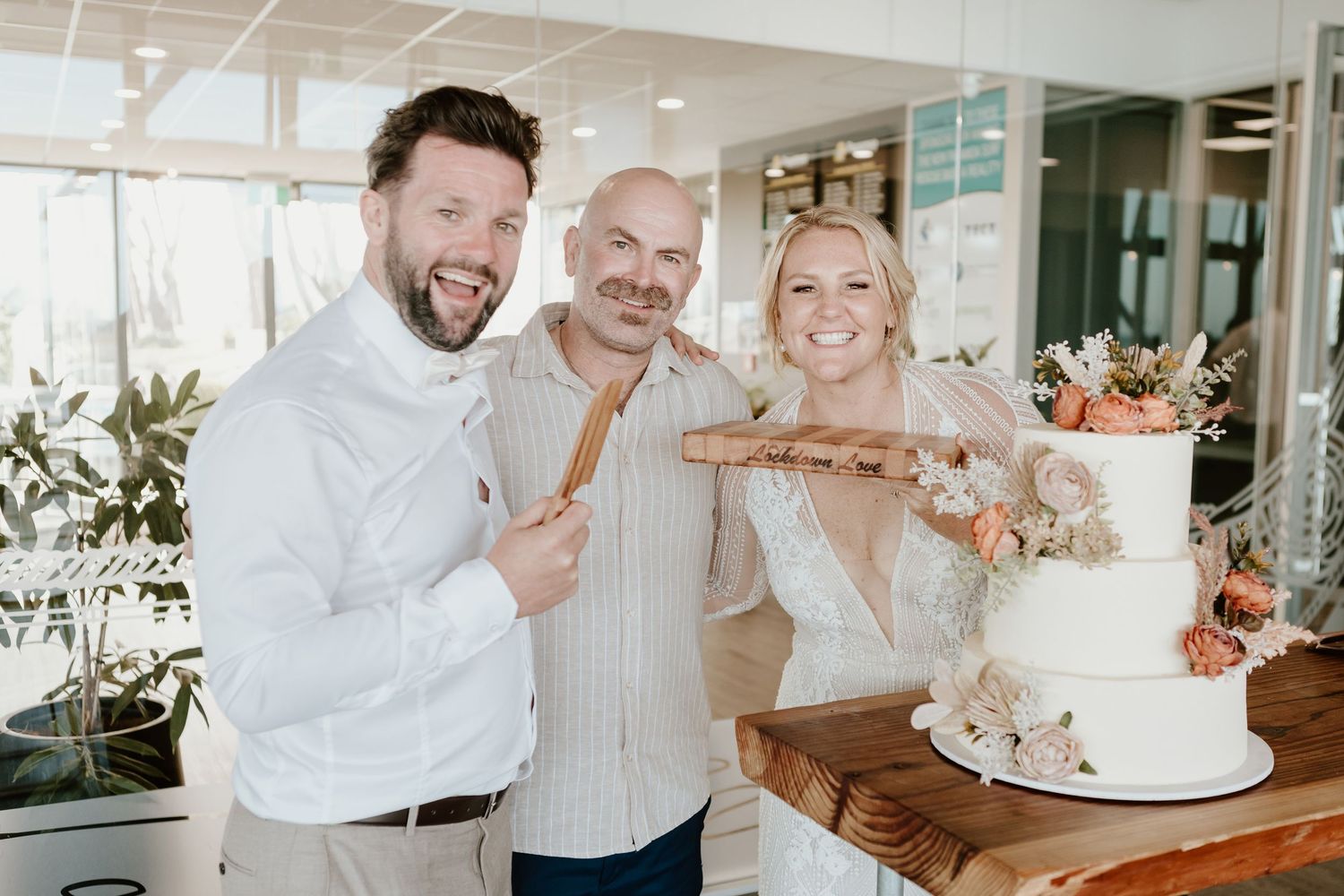 Three people laughing and posing with a three-tier white wedding cake at an indoor reception venue.