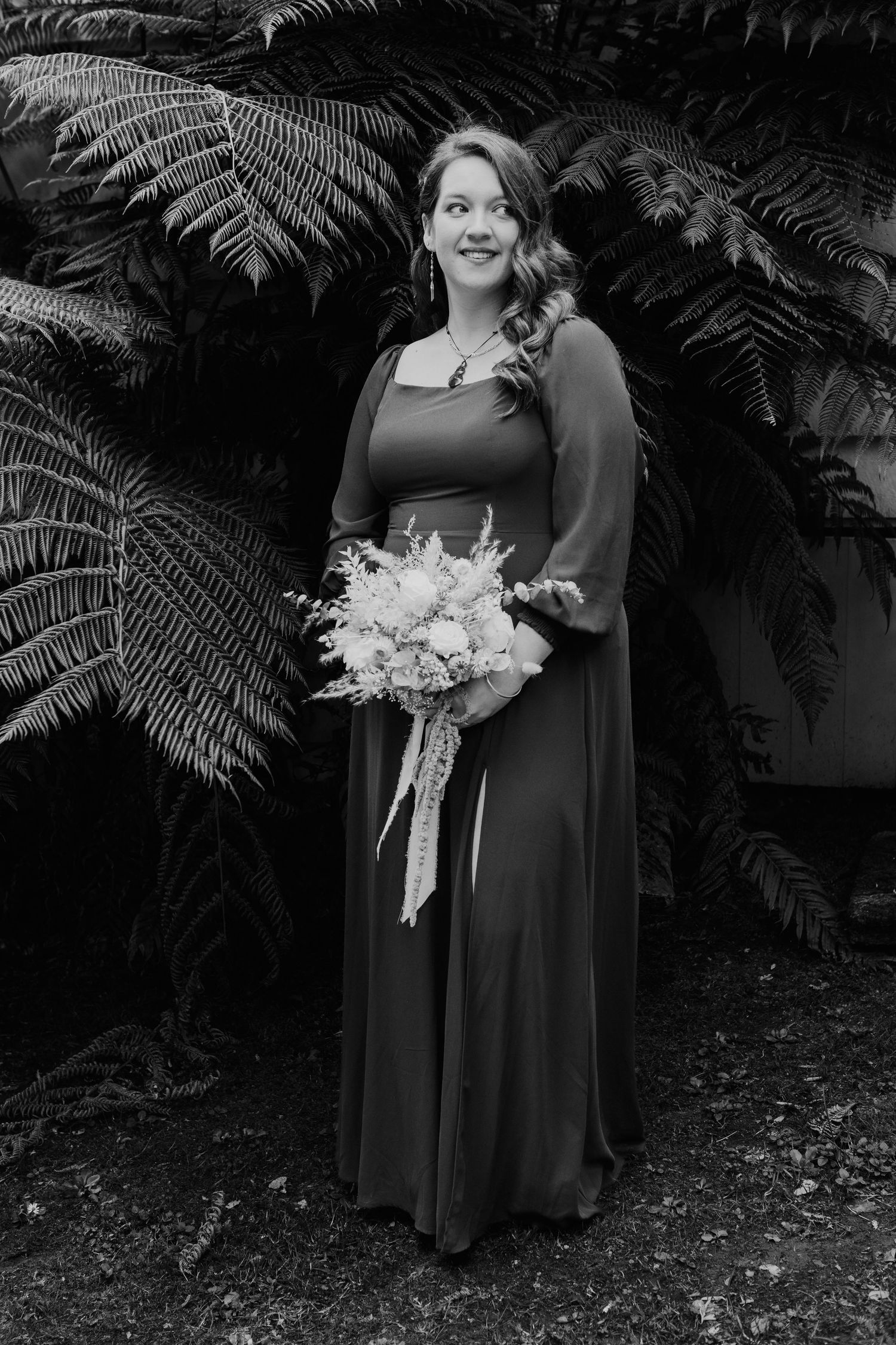 Black and white portrait of someone in a flowing dress holding a wedding bouquet, standing among lush ferns.