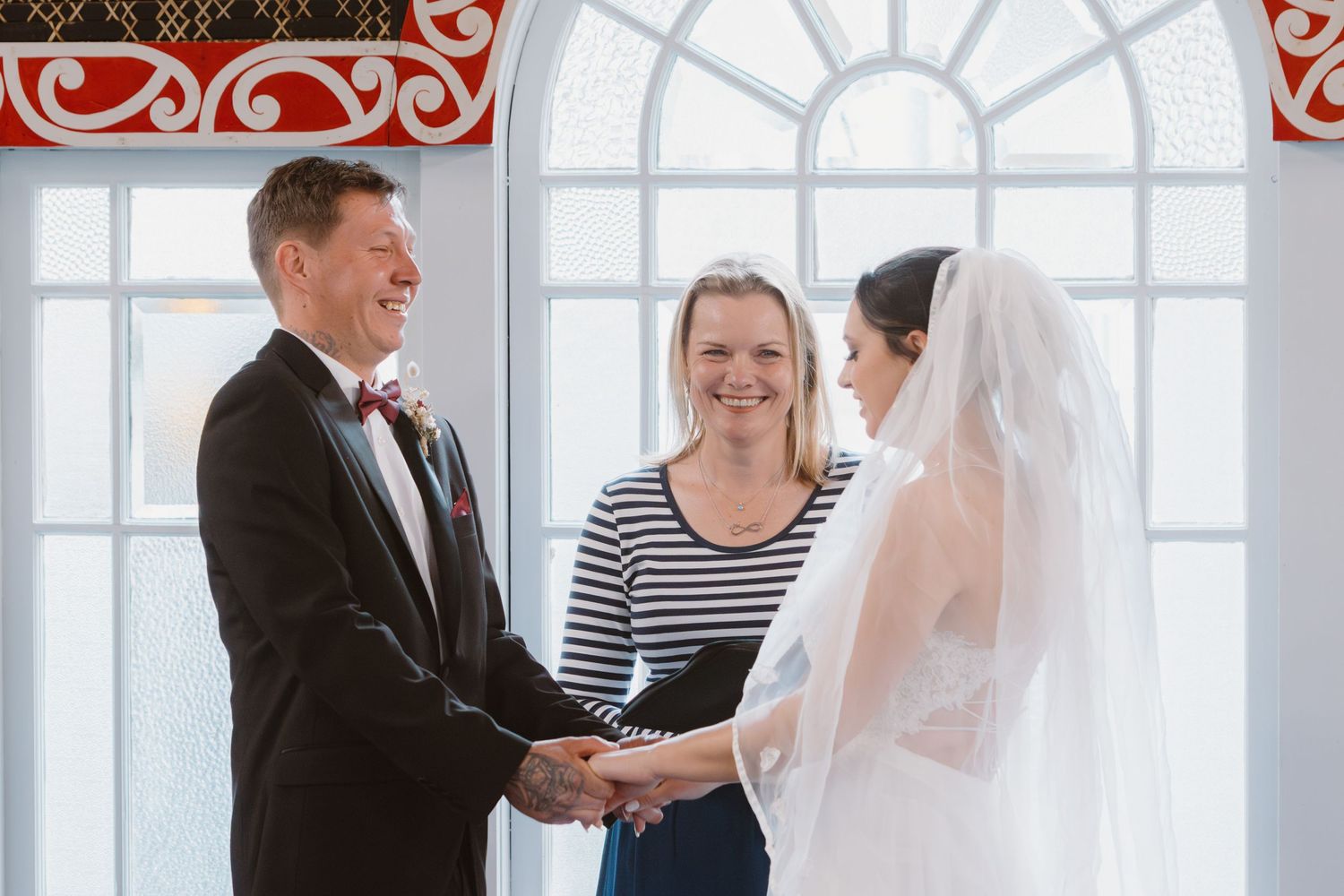 Wedding ceremony takes place under arched window with decorative red trim in wooden chapel.