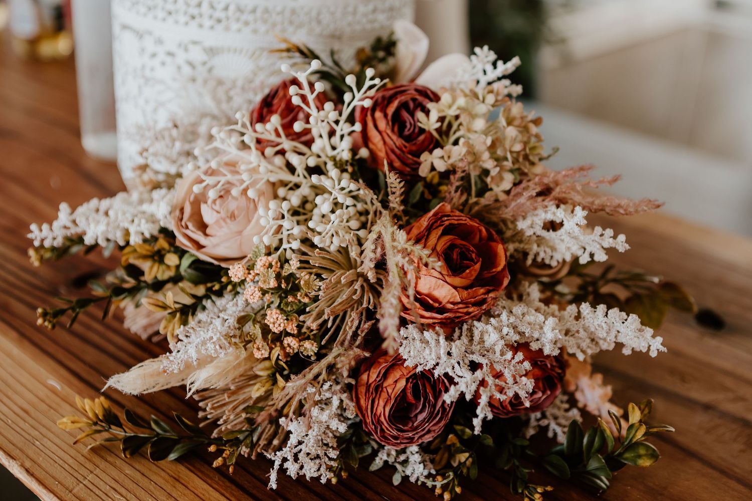 A dried floral bridal bouquet with burgundy and blush roses mixed with delicate white flowers on wooden surface. Tauranga wedding photograph