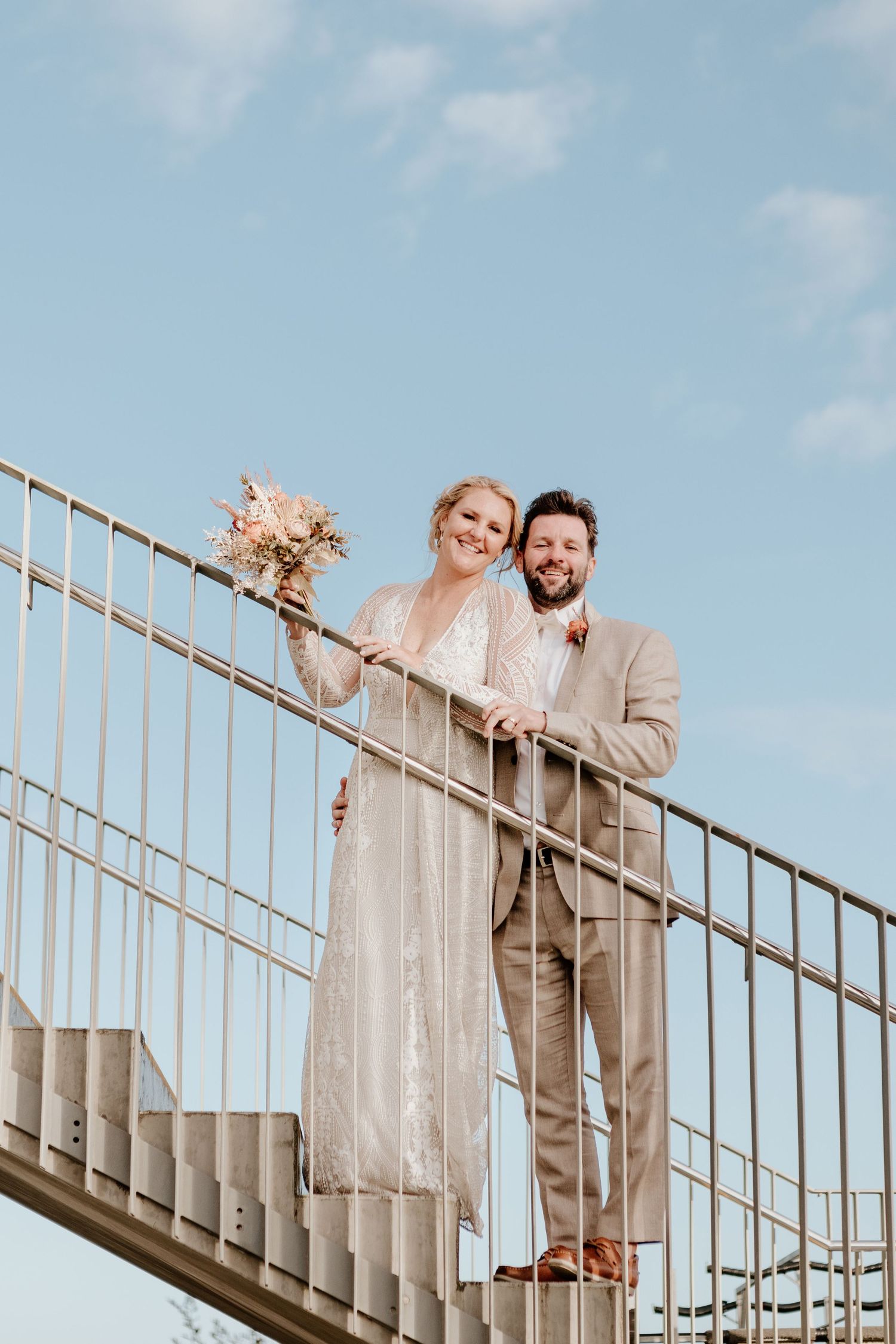 A couple poses on an outdoor staircase against a blue sky during their wedding celebration. Papamoa Surf Lifesaving Club Wedding
