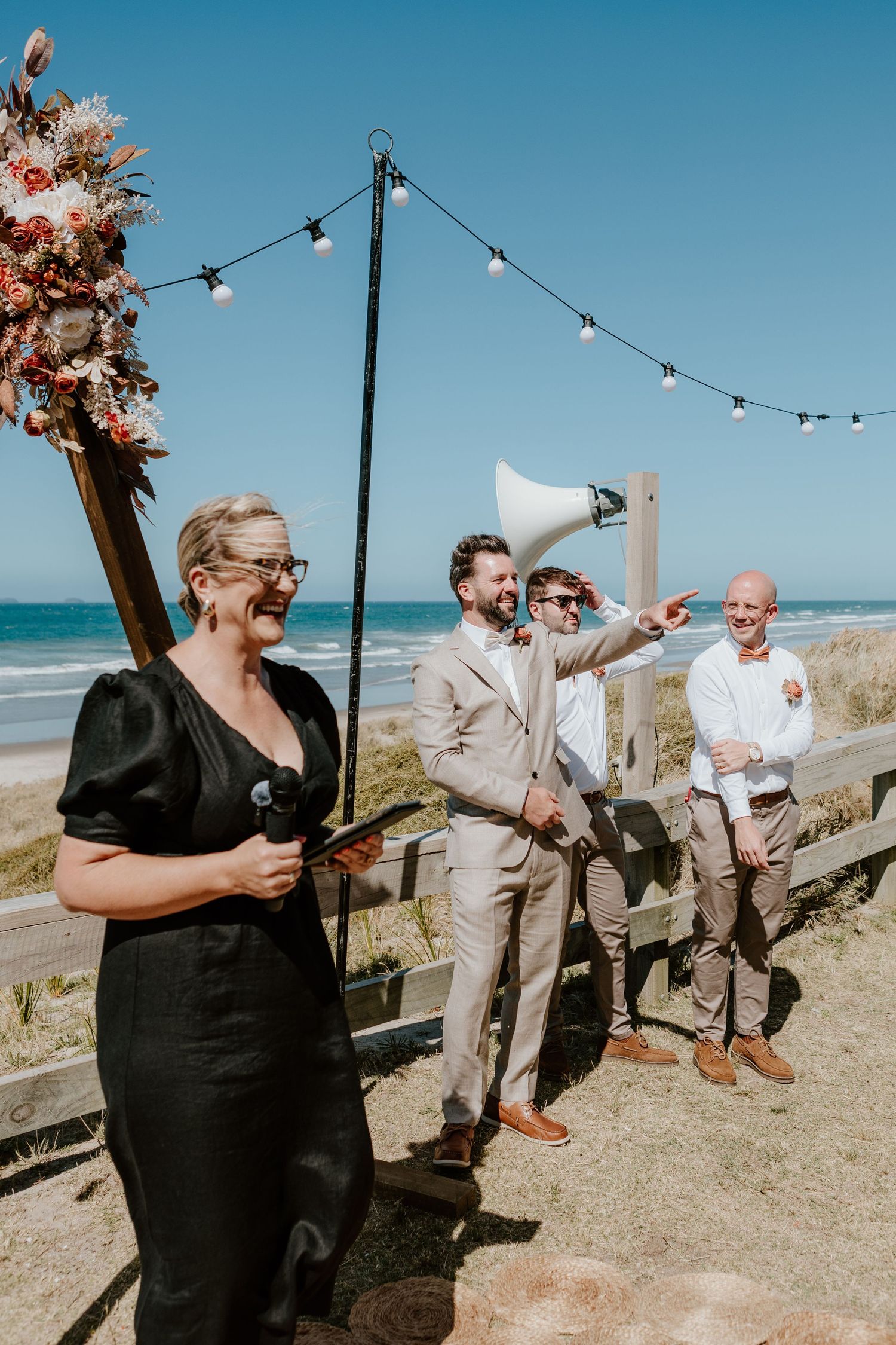 Guests in formal attire laugh and celebrate on a sunny beachfront deck with string lights. Papamoa Surf Lifesaving Club Wedding