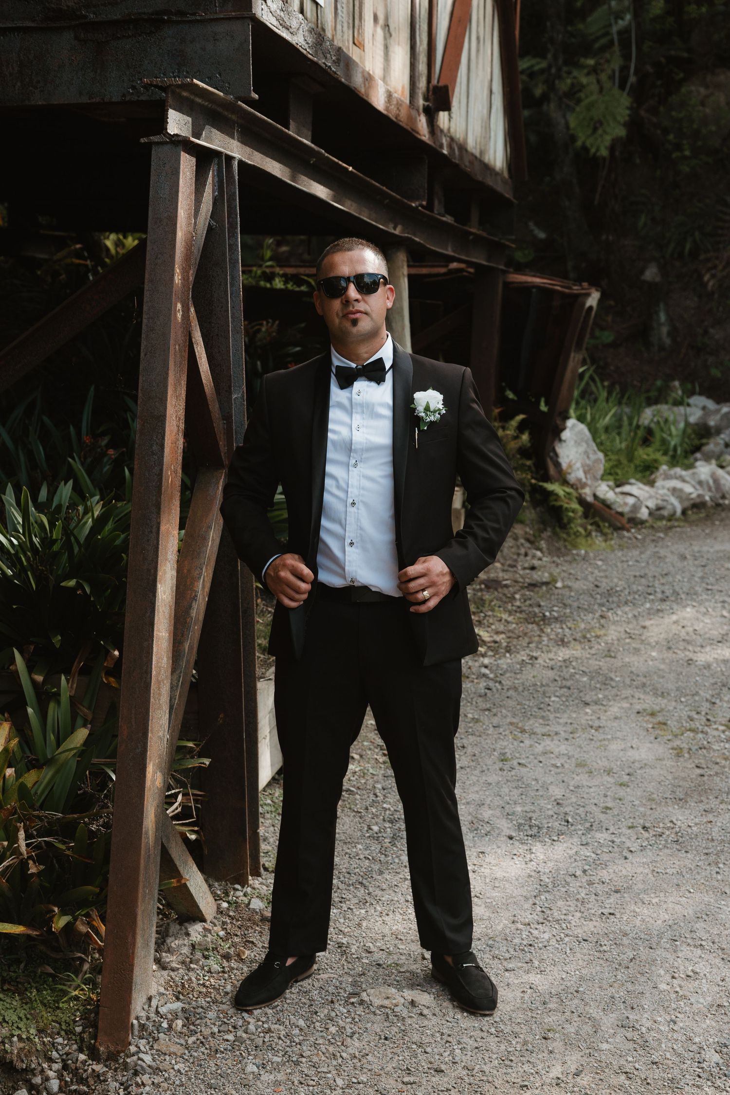 A person in a classic black tuxedo and sunglasses stands confidently on a gravel path near a rustic wooden structure.