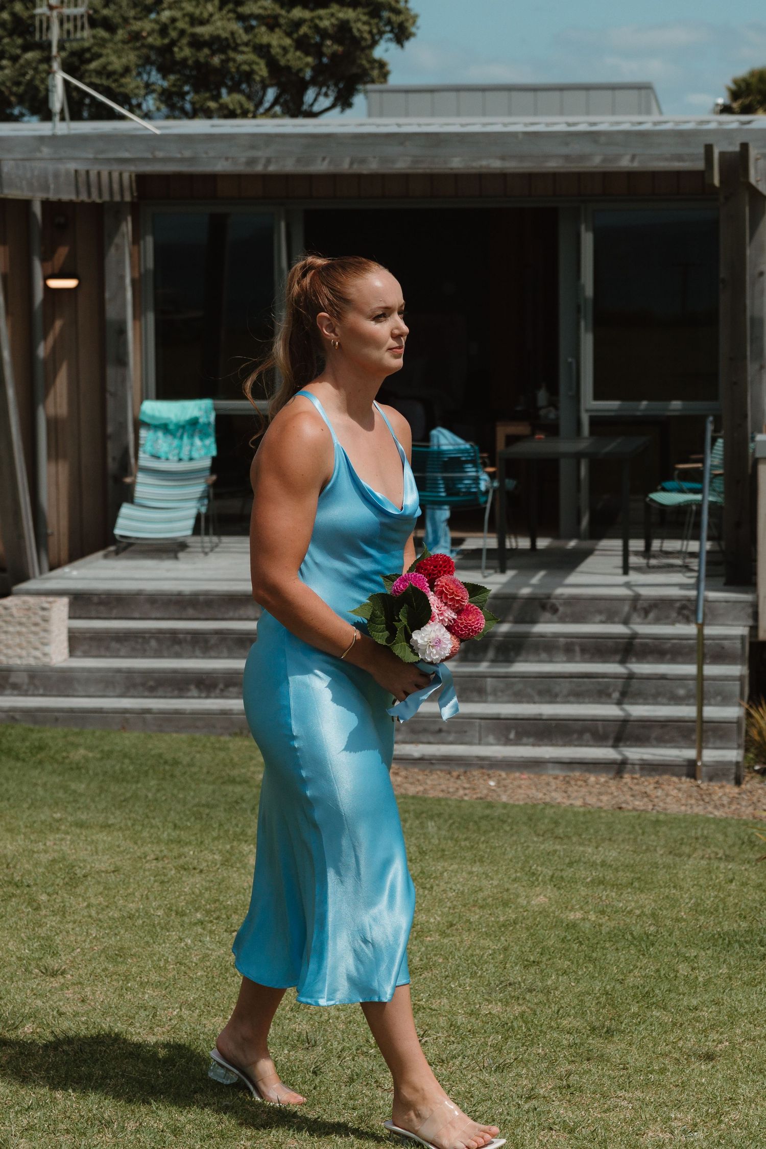 Woman in blue satin midi dress holding pink bouquet walks on green lawn outside modern beach house.