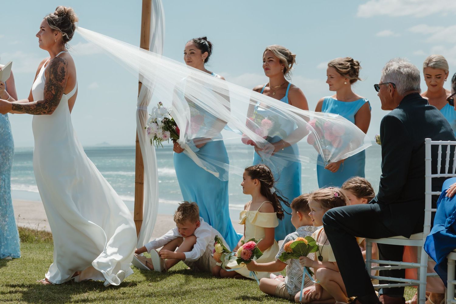 Bride in white gown with veil blowing in wind at outdoor beach wedding ceremony with bridesmaids in blue dresses.