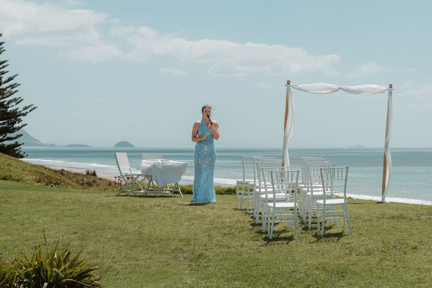 Outdoor beach wedding ceremony setup with white chairs, floral arch, and bridesmaid in blue dress by the ocean.