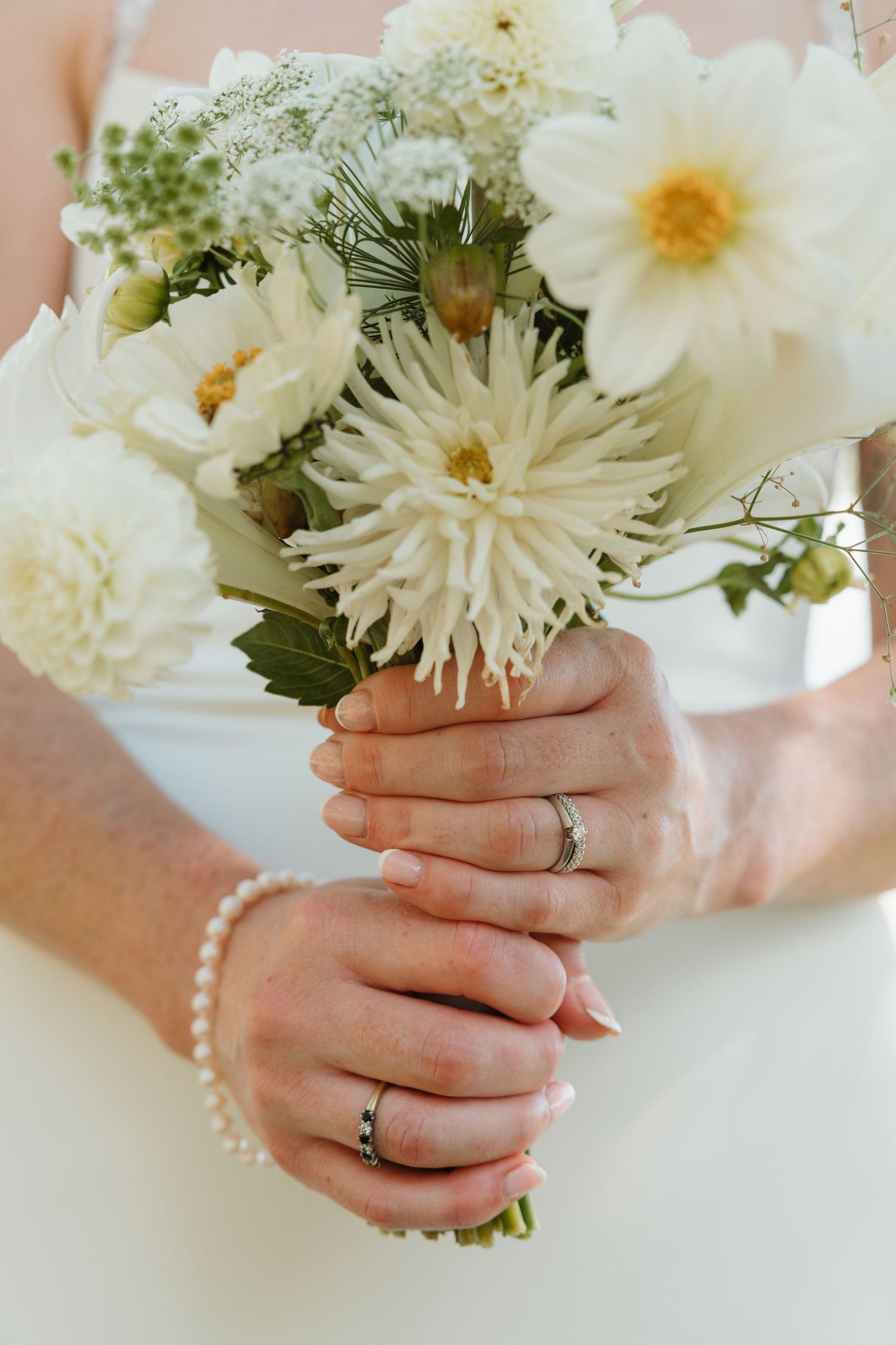 Bride holding white dahlia wedding bouquet with pearl bracelet and wedding ring on her hands.