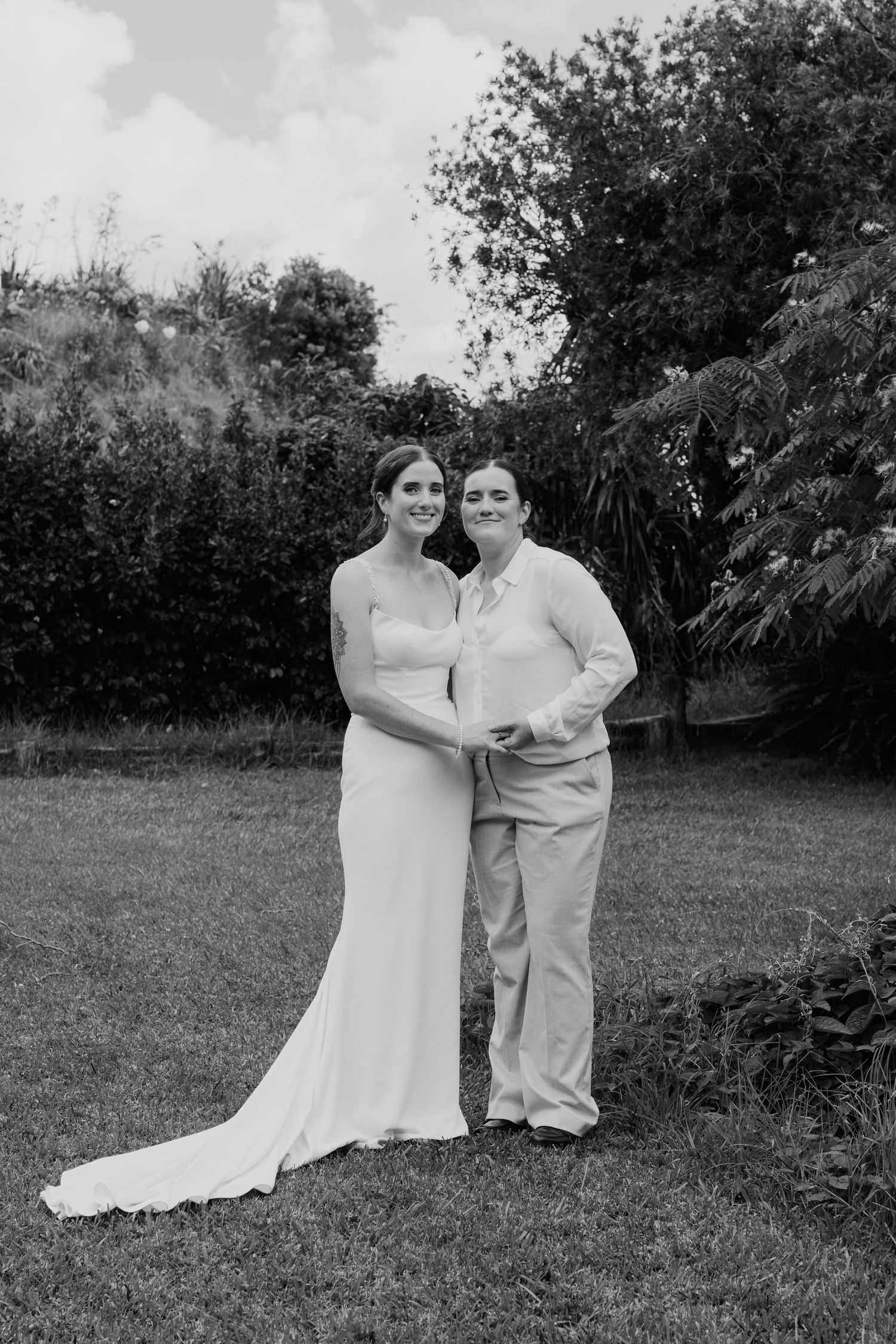 Black and white wedding photo of two brides posing together on a green lawn with trees in the background.