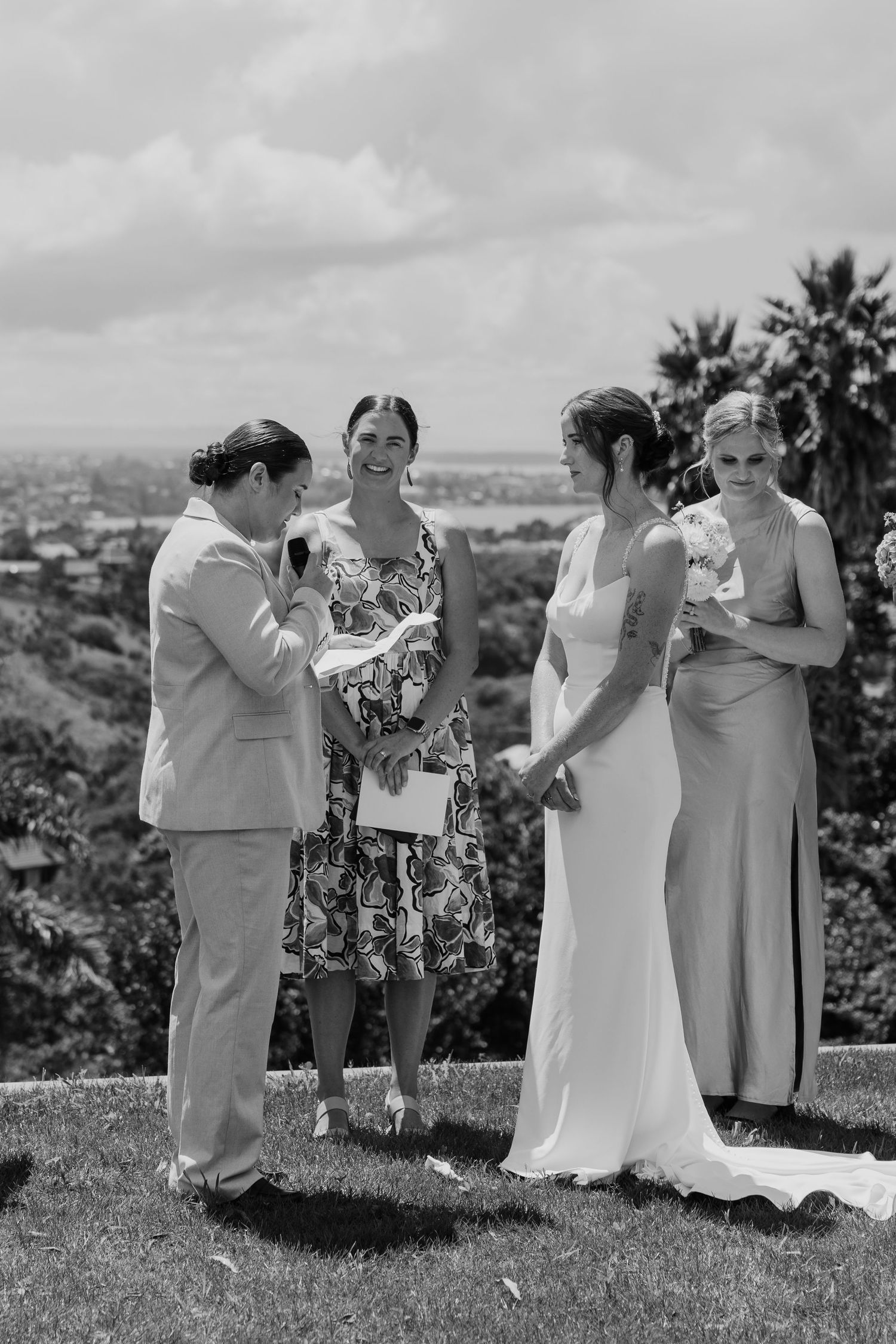Black and white outdoor wedding ceremony with two brides holding hands, officiant reading vows, and bridesmaid on sunny hilltop.