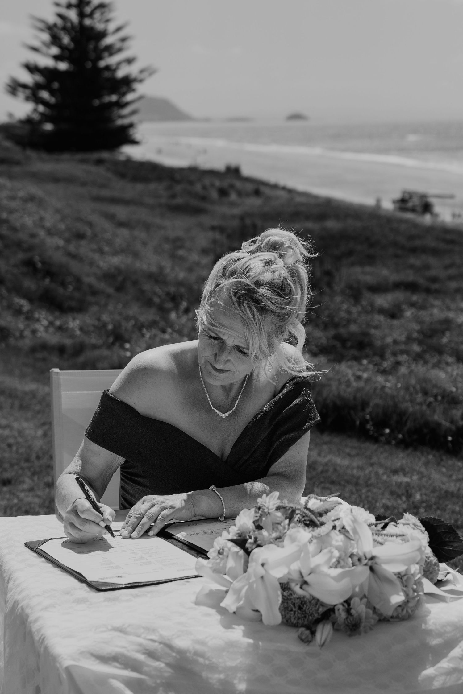 Black and white photo of a woman in an off-shoulder dress signing documents at a table with flowers, ocean backdrop.