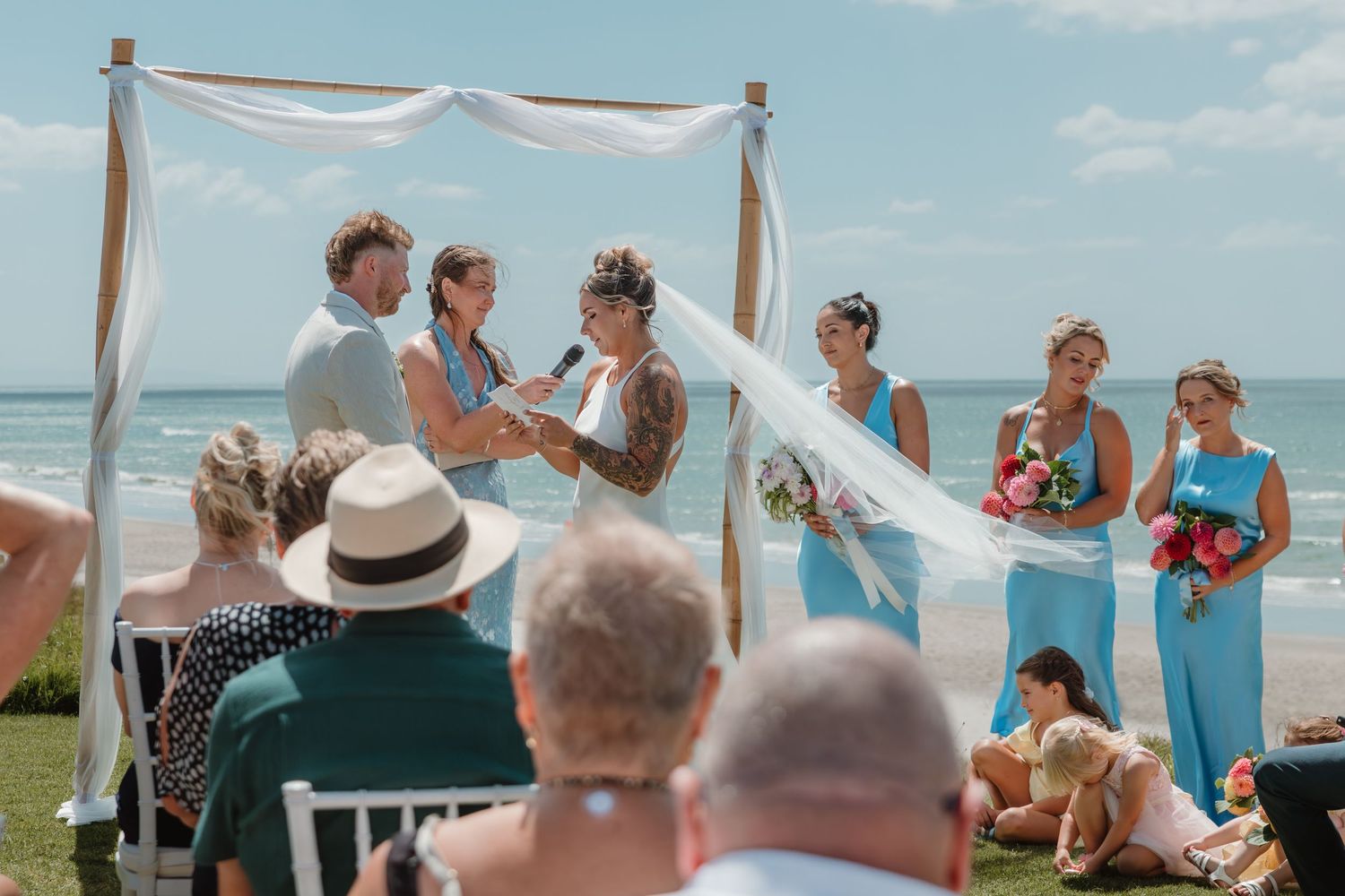 Outdoor beach wedding ceremony with couple exchanging vows under a wooden arch with white draping and bridesmaids in blue.