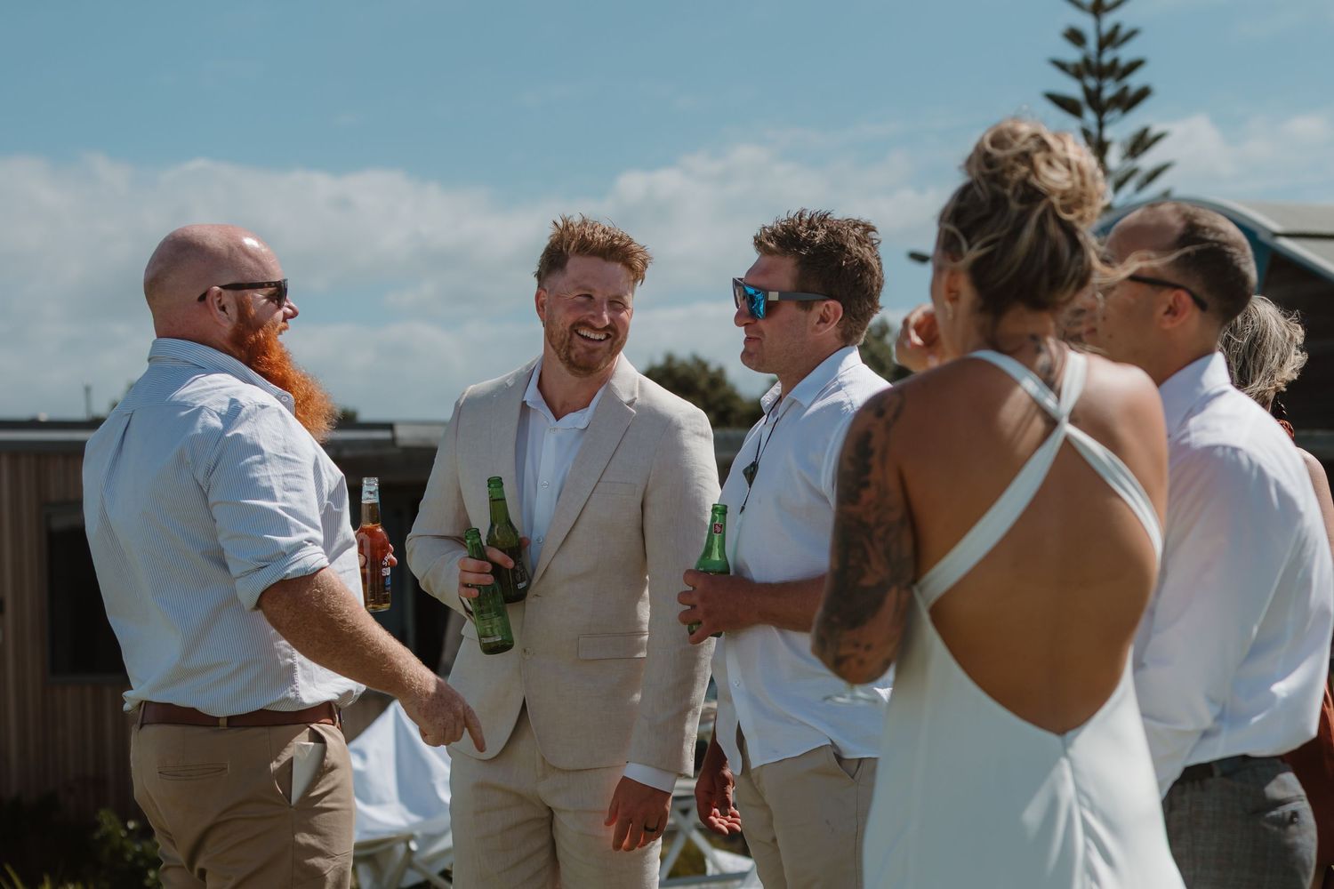 Wedding guests socializing outdoors, holding beer bottles, dressed in formal attire on a sunny day.