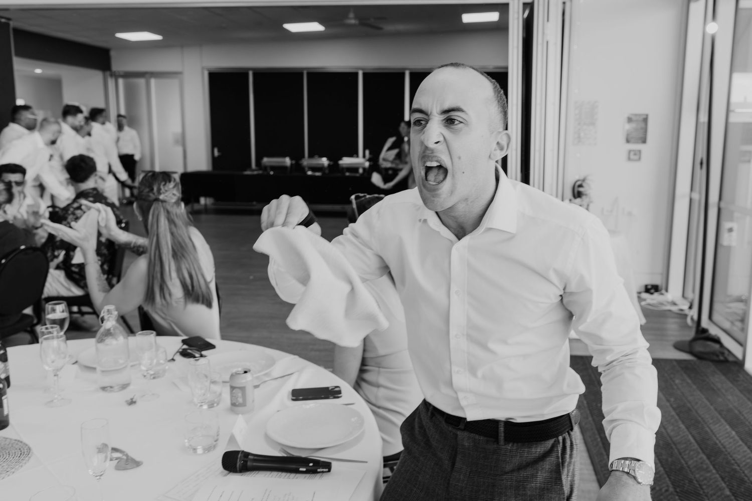 A man in a white shirt holds a microphone and food at a wedding reception dinner.