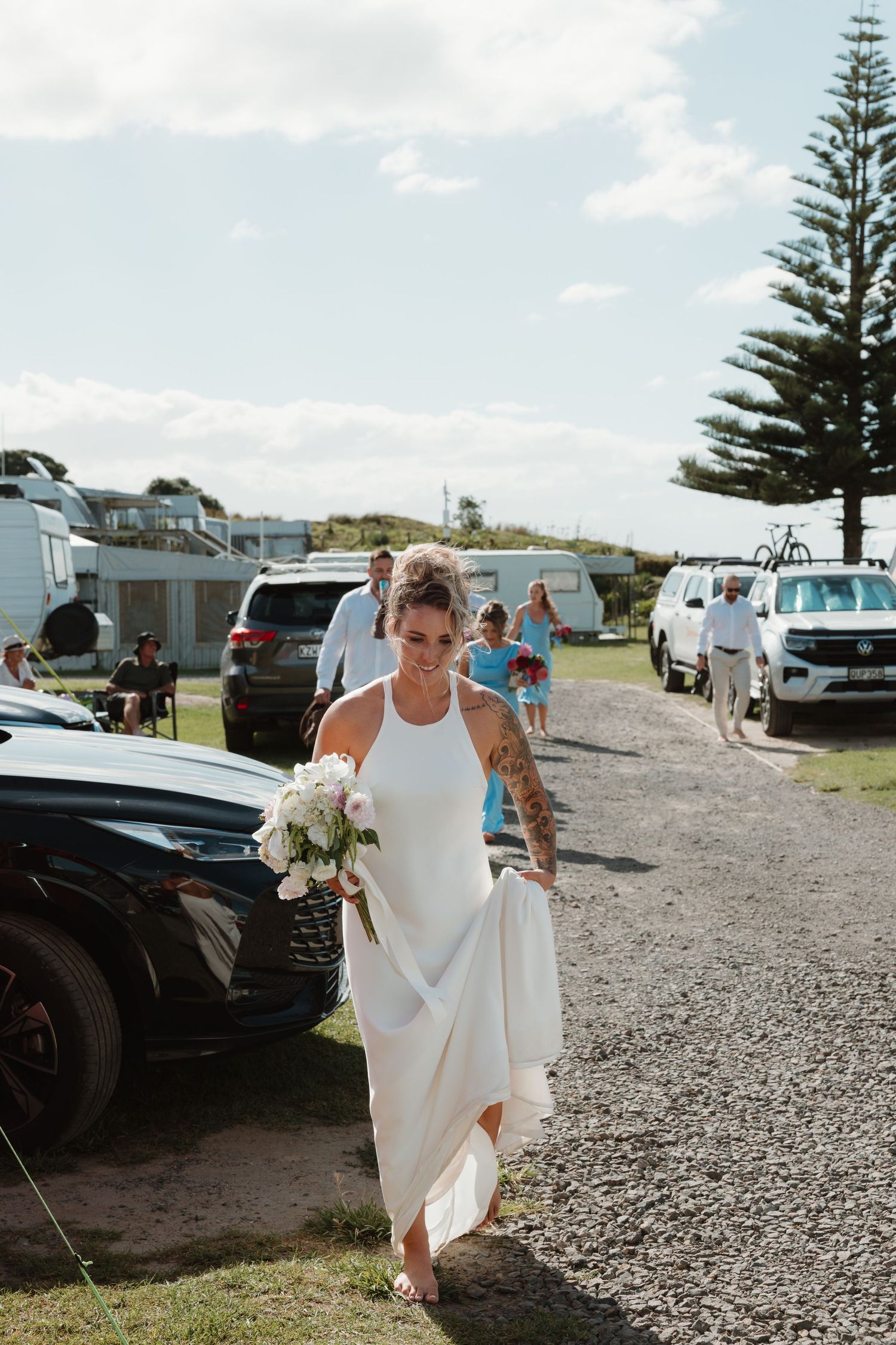 Barefoot bride in white halter gown holding bouquet walks through coastal campground on her wedding day.
