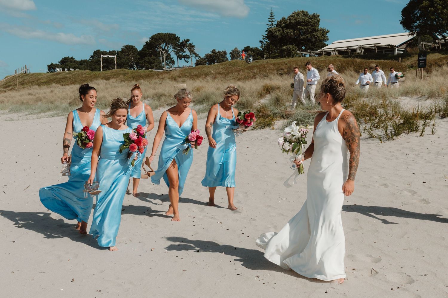 Bride in white gown walks toward bridesmaids in blue satin dresses on a sandy beach wedding ceremony.