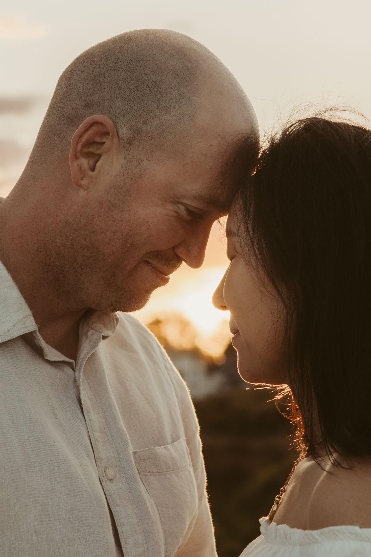 Expecting couple touch foreheads at sunset with sun flare and warm golden light creating a dreamy atmosphere. Tauranga maternity photoshoot
