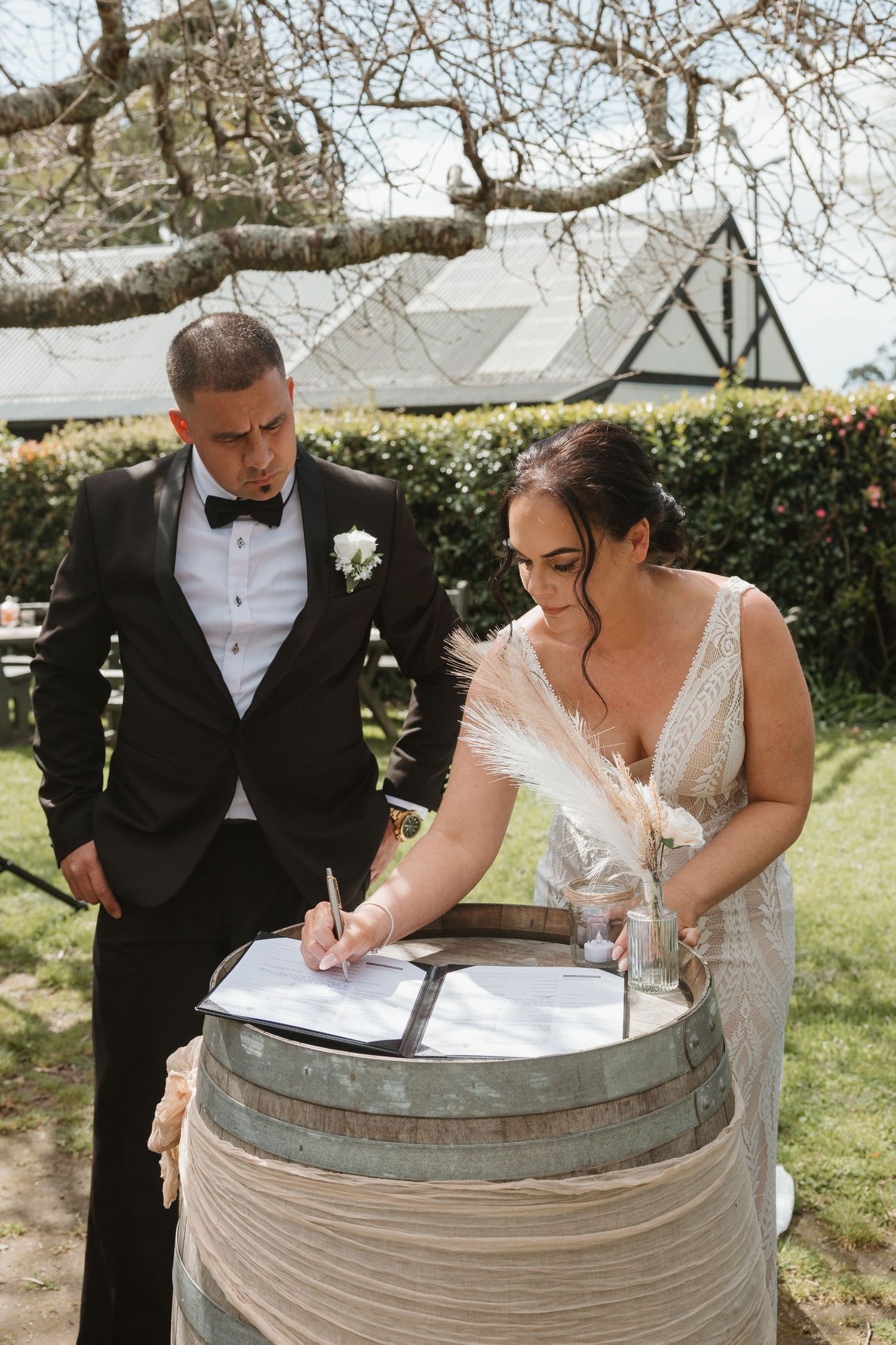 A couple in formal wedding attire signs their marriage license on a rustic wine barrel outdoors.