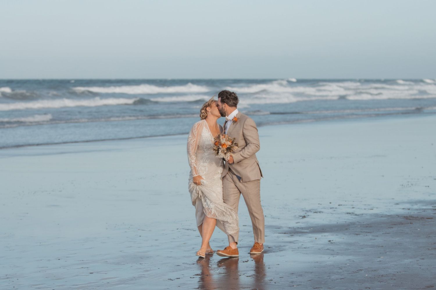 A couple in formal attire share an intimate moment while standing barefoot on a misty beach at sunset.