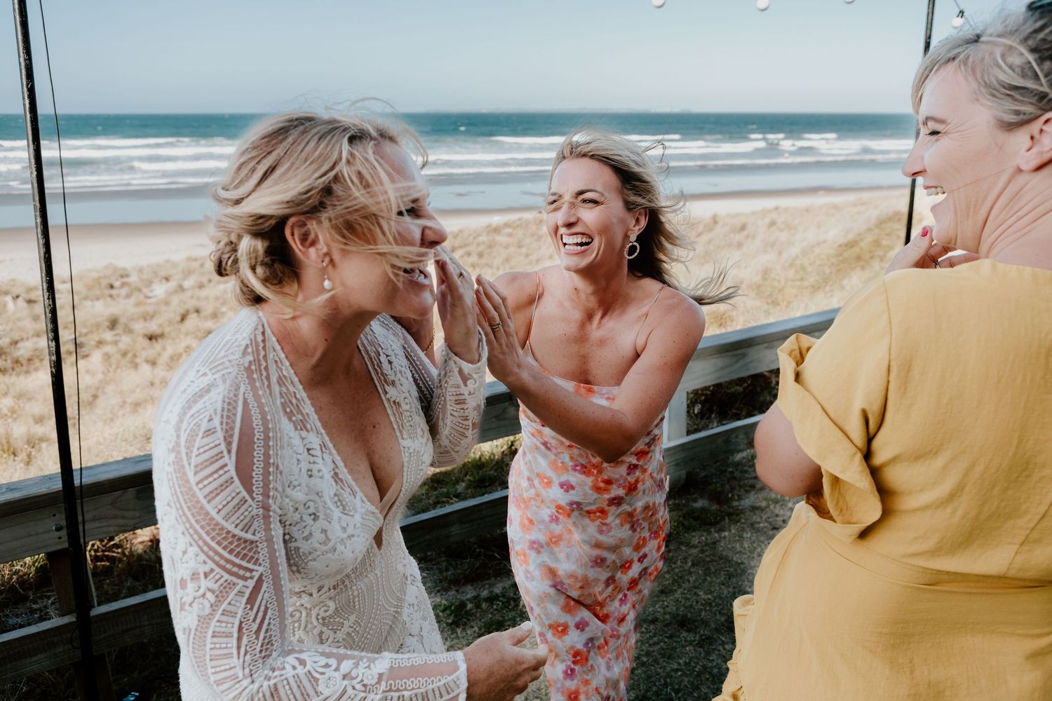Three friends sharing laughter and joy together on a sunny day at the beach with ocean waves in the background.