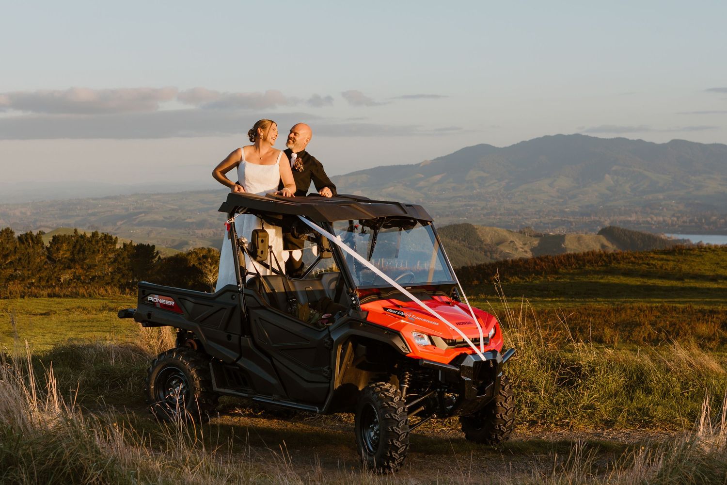 Two people lean against a red ATV side-by-side vehicle on a hilltop overlooking scenic mountain views at sunset.