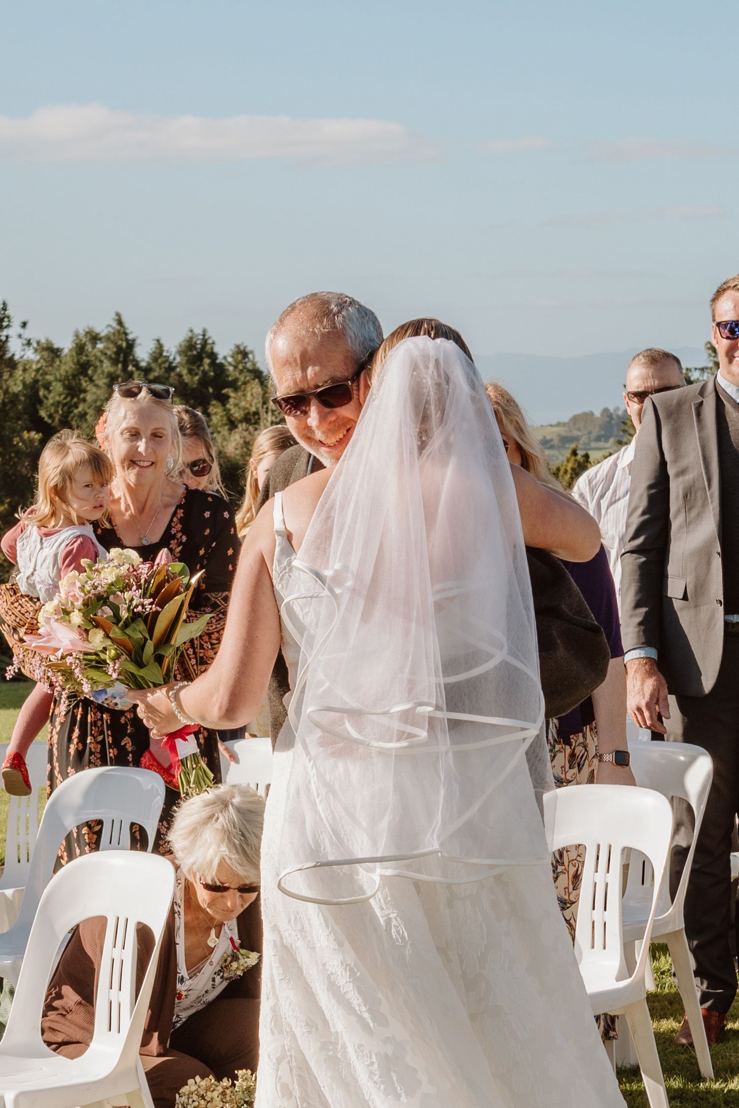 An outdoor wedding ceremony with guests seated in white chairs as a bride in a flowing white dress and veil greets attendees.