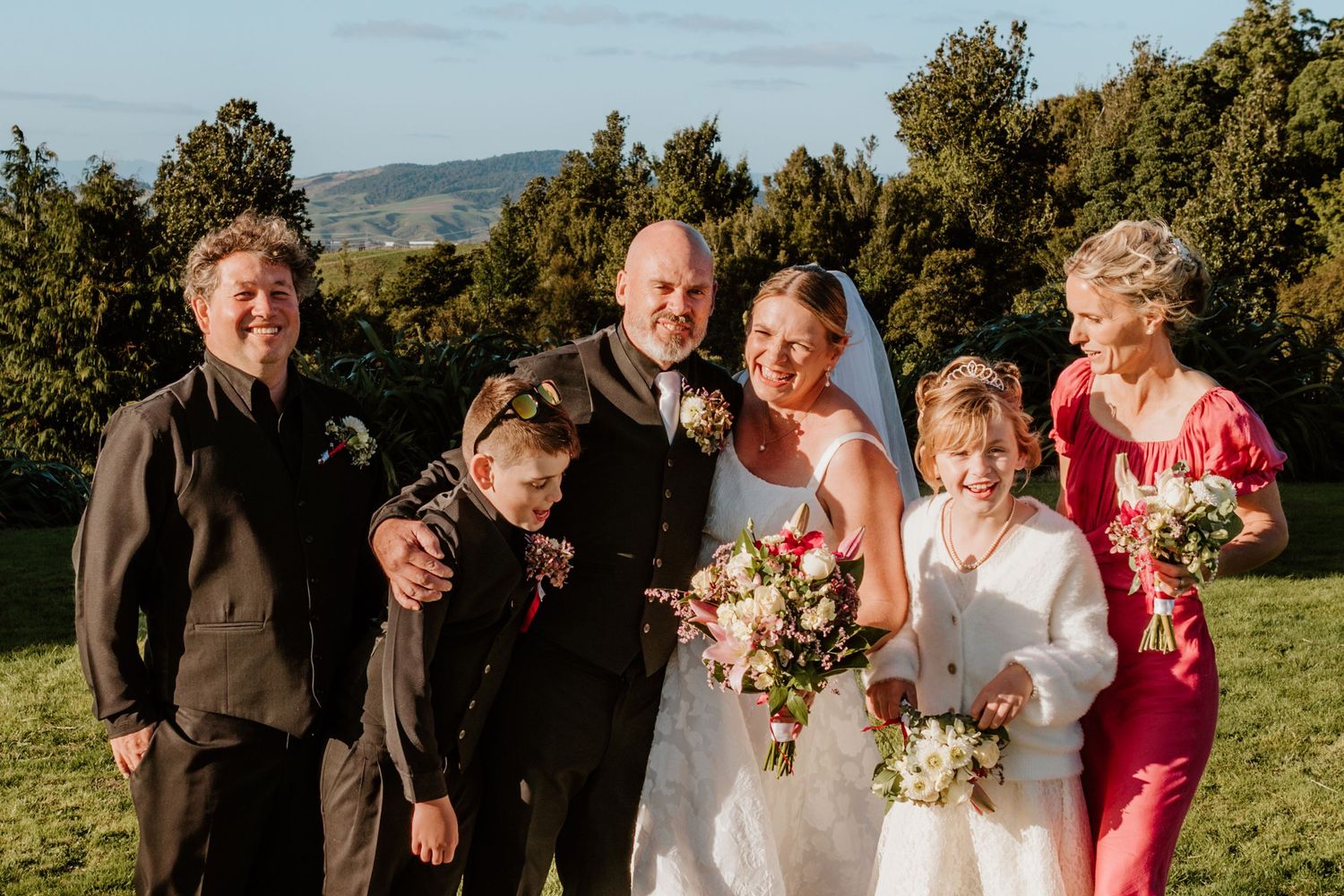 Wedding party poses together on grassy hilltop with lush green trees and mountains in background.