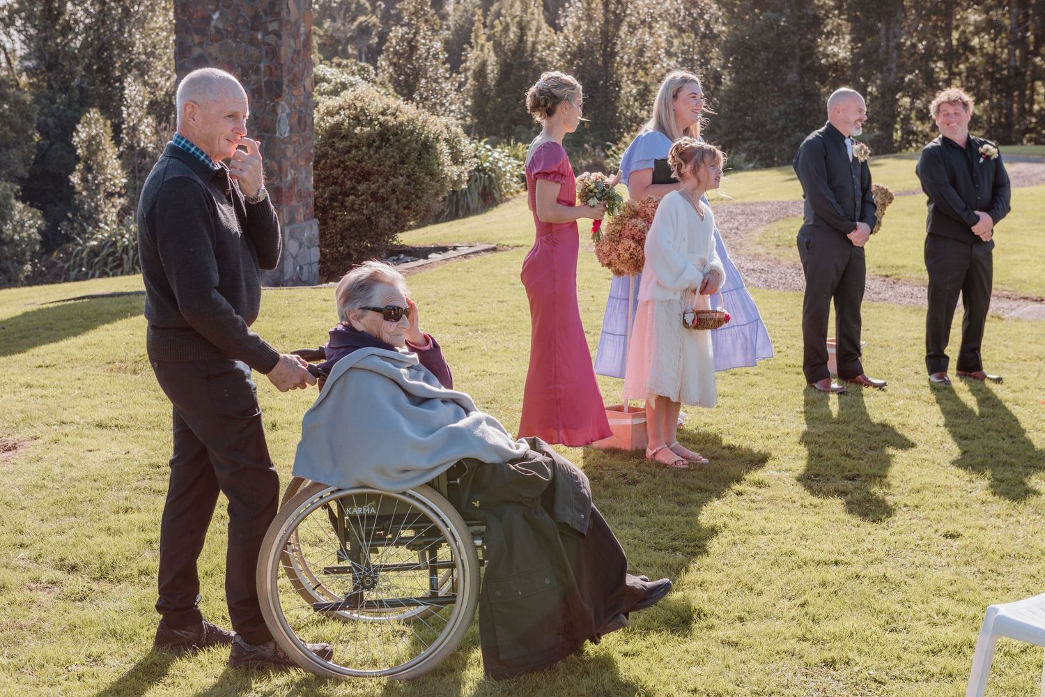 Wedding guests gather on a sunny lawn with one person seated in a wheelchair while others stand in formal attire.
