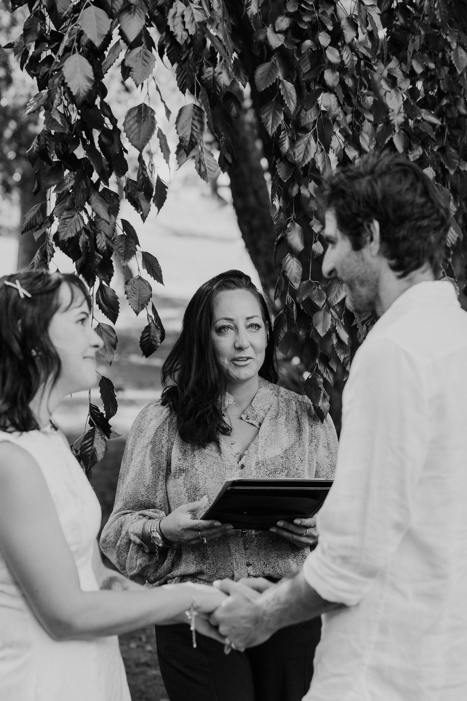 Black and white outdoor wedding ceremony with couple holding hands before officiant under leafy tree.