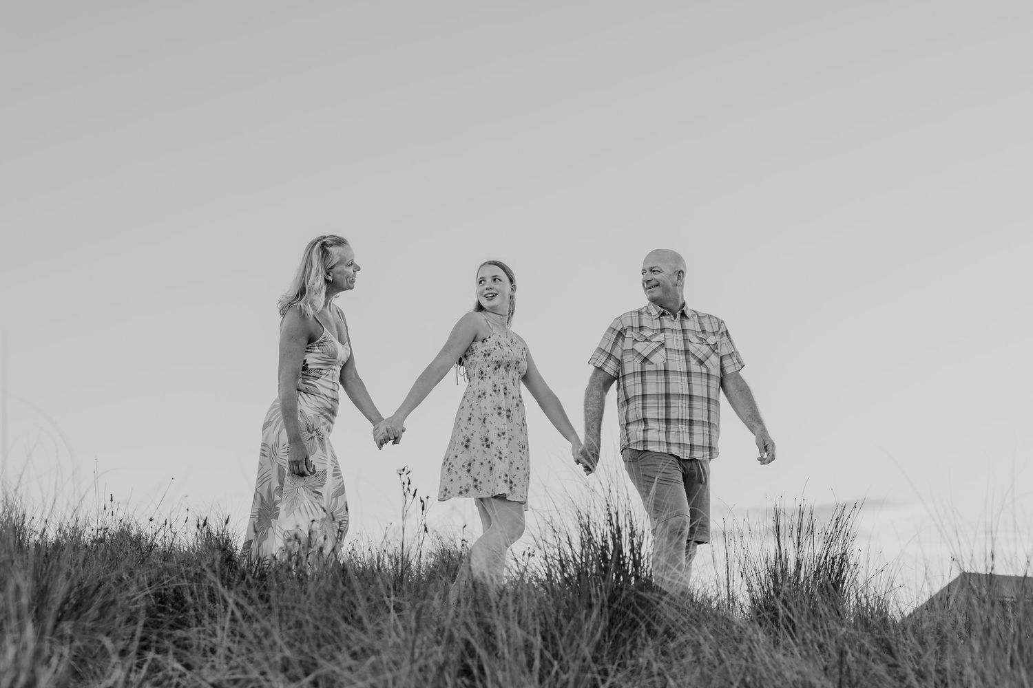 Black and white photo of a family of three holding hands and walking through tall grass outdoors.