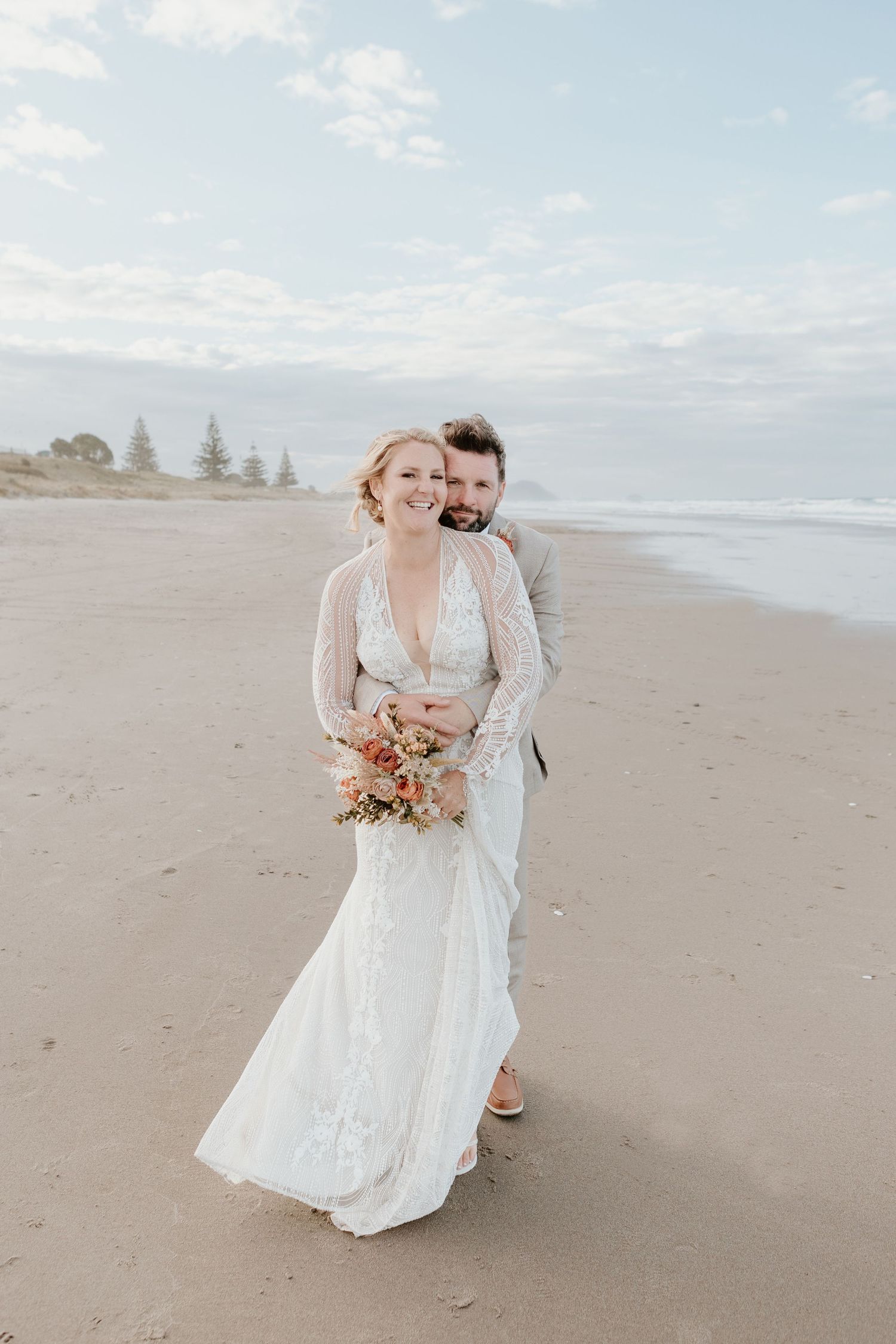 A happy couple in wedding attire embraces while standing on a peaceful beach with pine trees in the background.