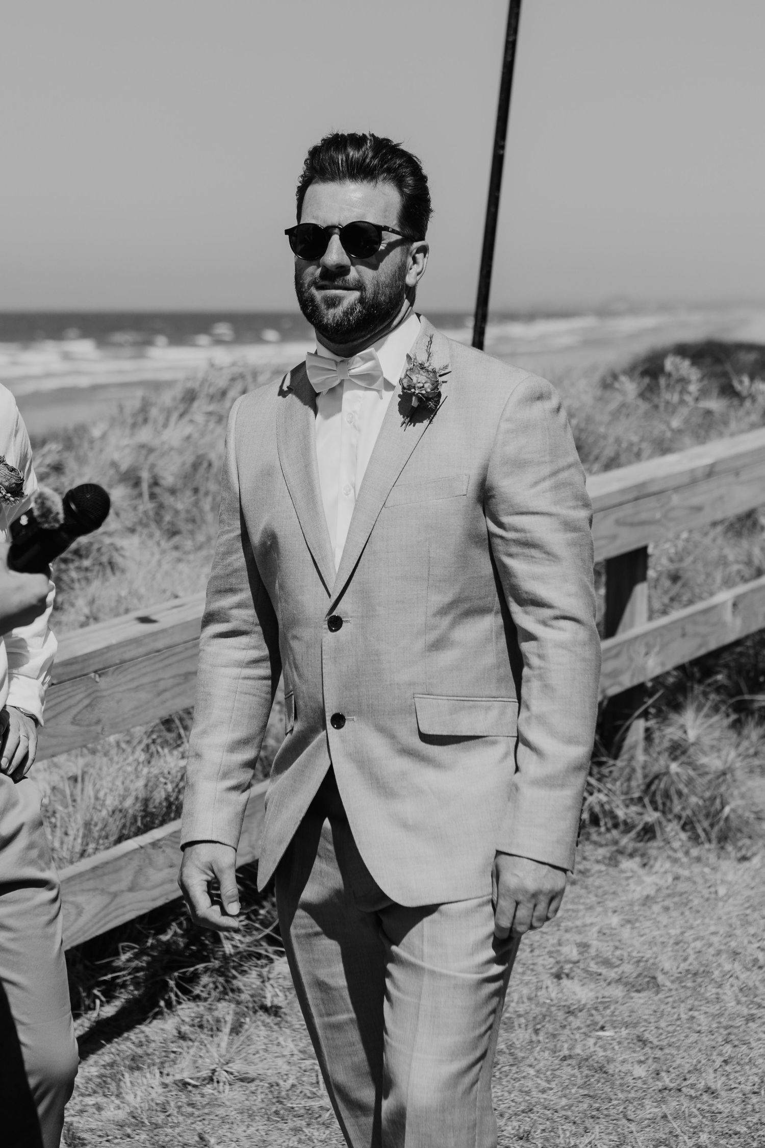 A groom in a light gray suit and sunglasses smiles at a beach wedding ceremony with ocean views in black and white.