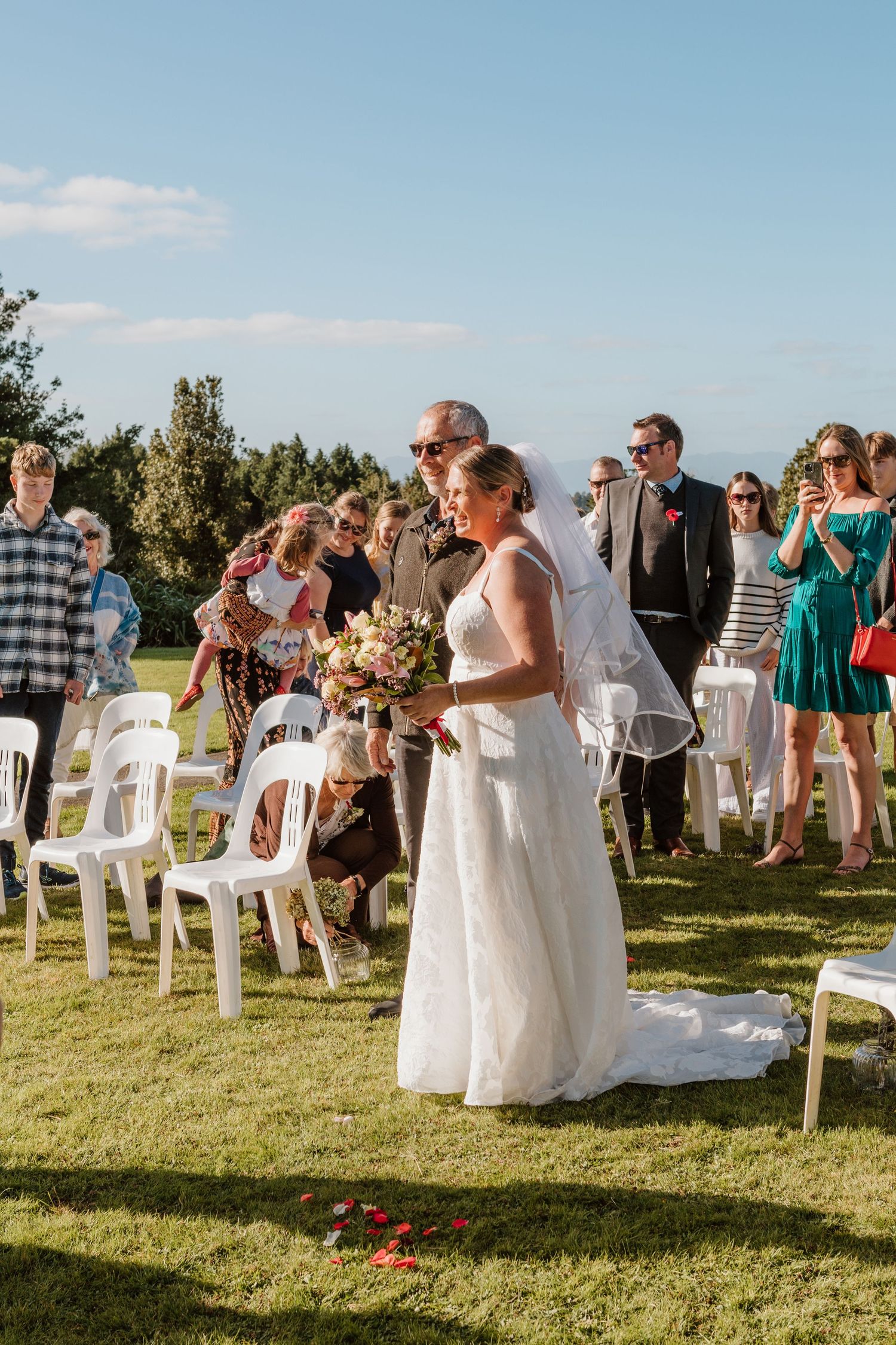An outdoor wedding ceremony takes place on a grassy lawn with white chairs and guests gathered under blue summer skies.