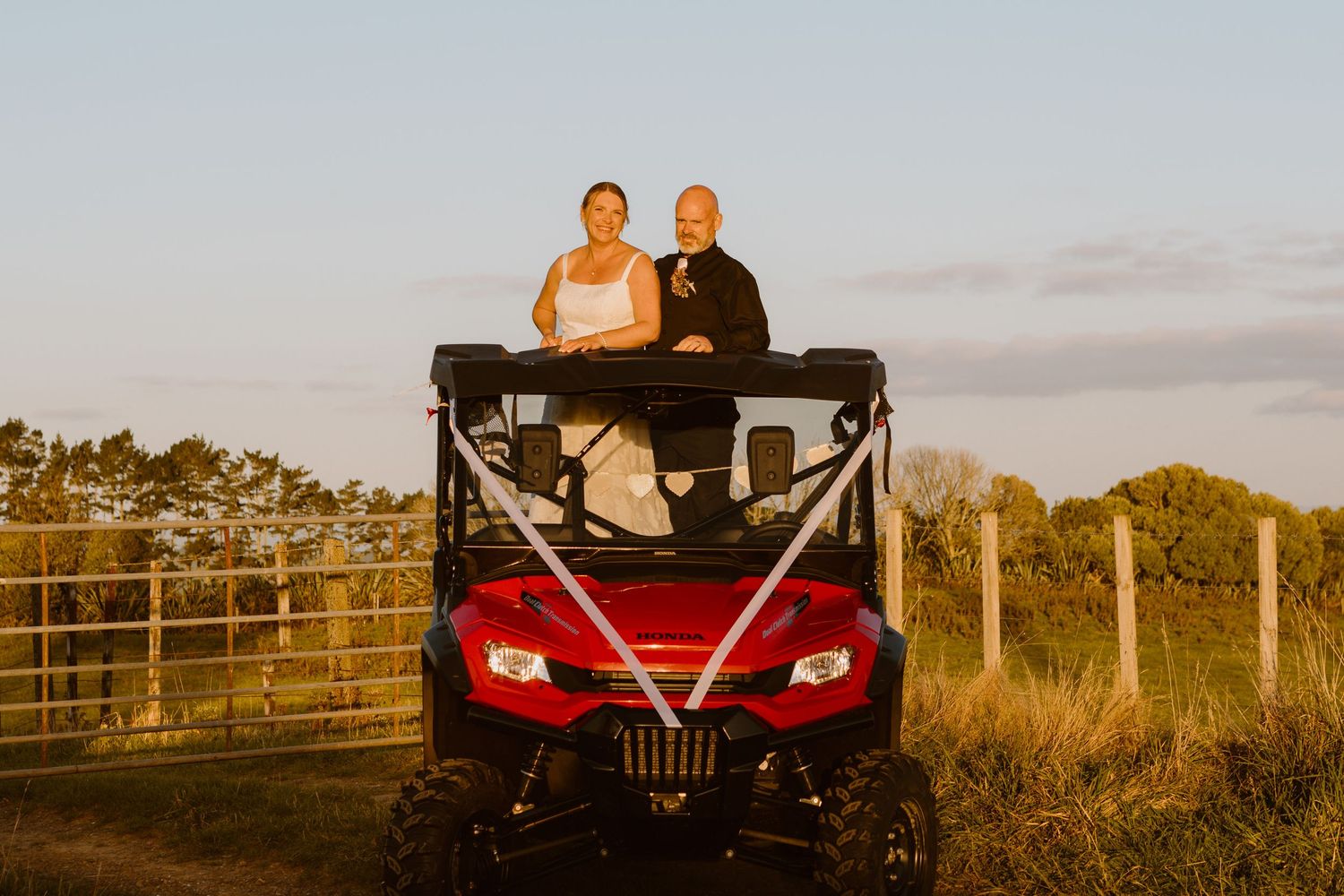 A couple sits atop a red utility vehicle decorated with ribbons at sunset in a rural setting with fence posts visible.