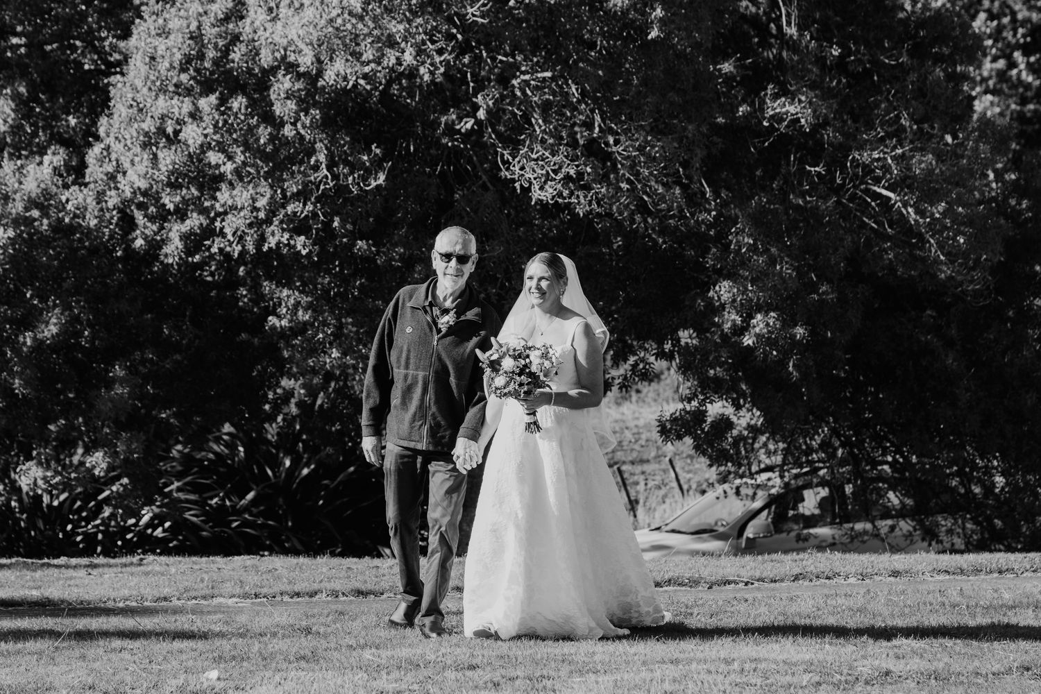 A black and white photo captures a wedding couple on their special day standing outdoors beneath lush trees.